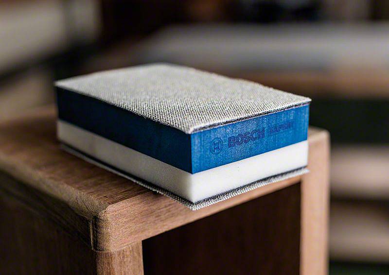 Niebieski gąbkowy papier ścierny Bosch leży na krawędzi drewna. Idealny do precyzyjnego szlifowania w narożnikach i trudno dostępnych miejscach.
