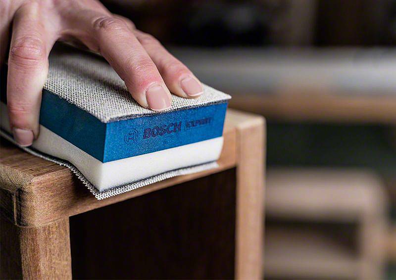 Ręka szlifuje drewnianą powierzchnię blokiem szlifierskim. Blok szlifierski jest niebieski z nadrukiem 'BOSCH Expert'.