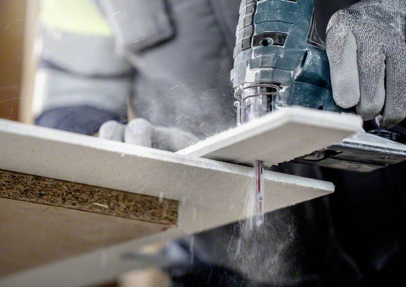 Osoba przecina drewnianą płytę elektryczną wyrzynarką. Podczas obróbki materiału leci pył i wióry.