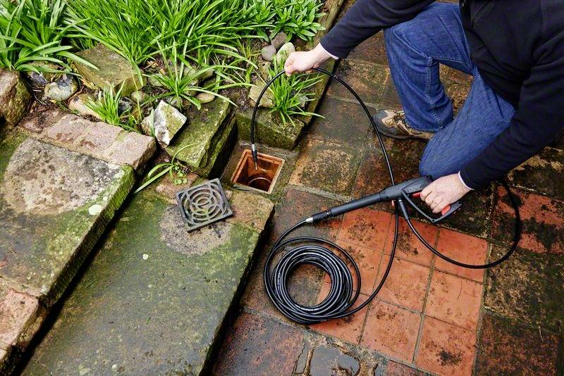 Osoba czyści zatkany odpływ w ogrodzie za pomocą węża wysokociśnieniowego. Widoczne są rośliny i mokre brukowane podłoże.