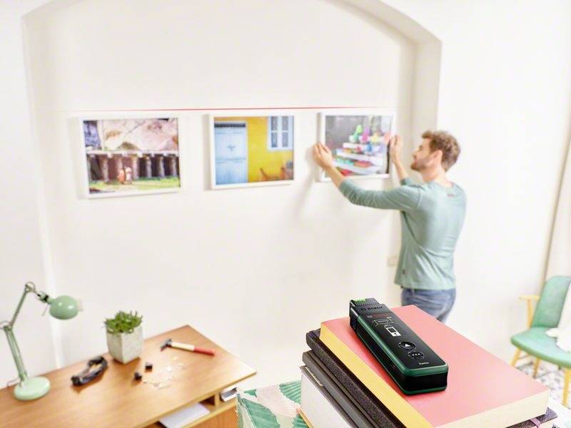 Mężczyzna zawiesza zdjęcia za pomocą narzędzia laserowego na białej ścianie. Na pierwszym planie leżą książki z urządzeniem laserowym na wierzchu.