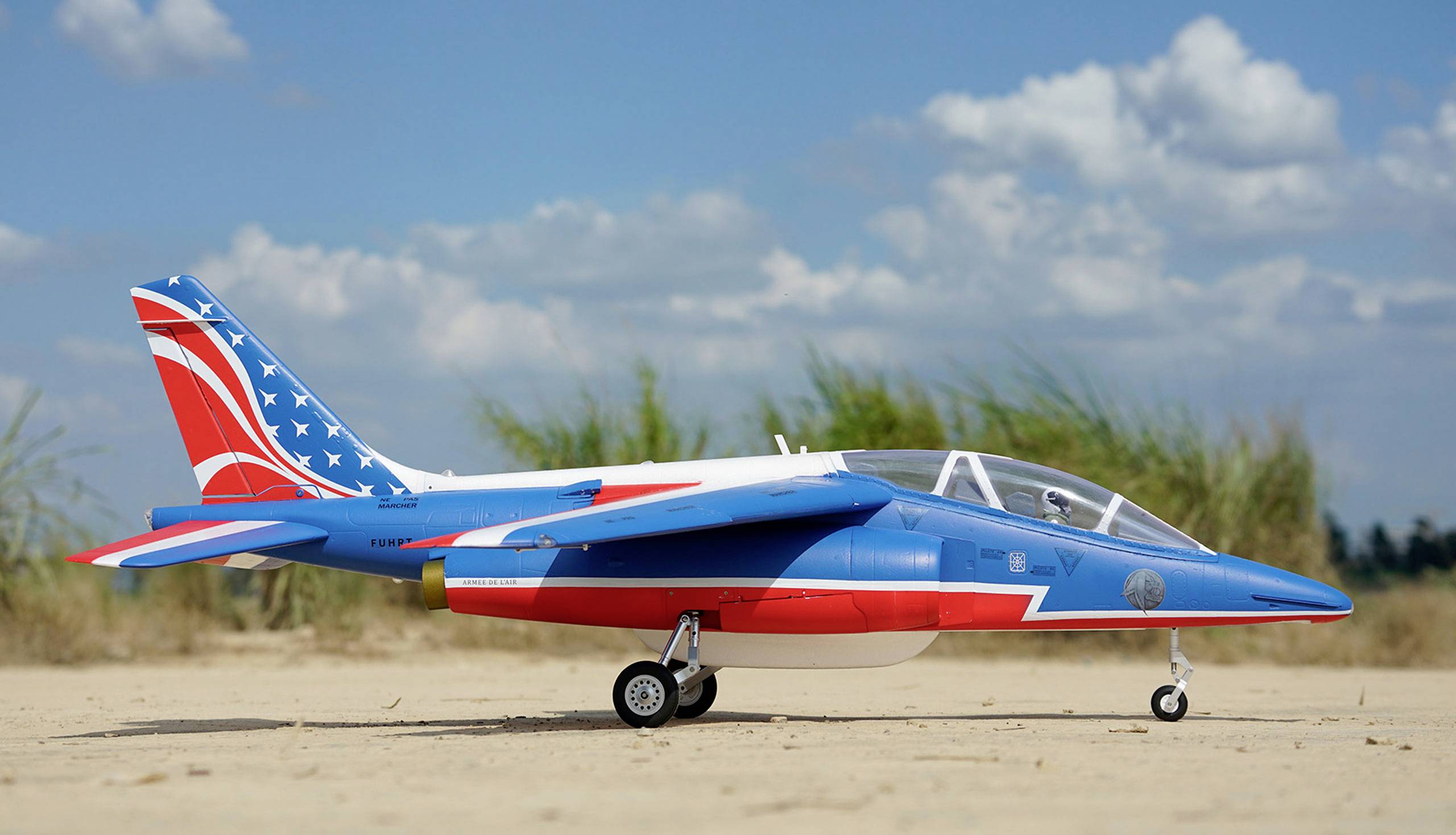 Kolorowo pomalowany model samolotu stoi na piaszczystym gruncie, z błękitnym niebem i kilkoma chmurami w tle.