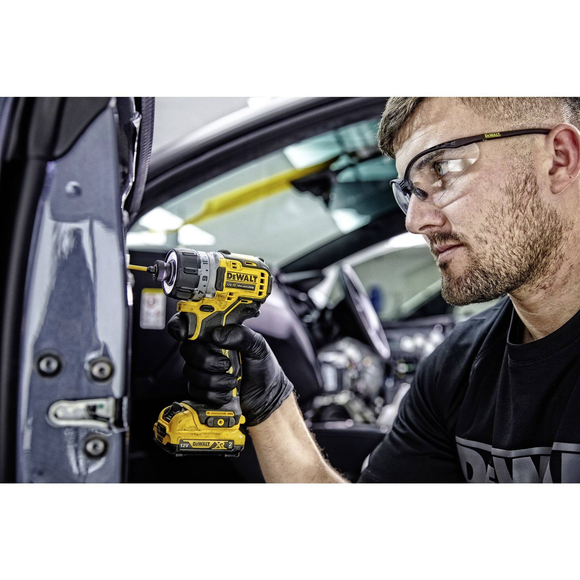 Osoba naprawia samochód za pomocą elektrycznego wkrętaka. Nosi okulary ochronne i rękawice. Samochód jest otwarty.