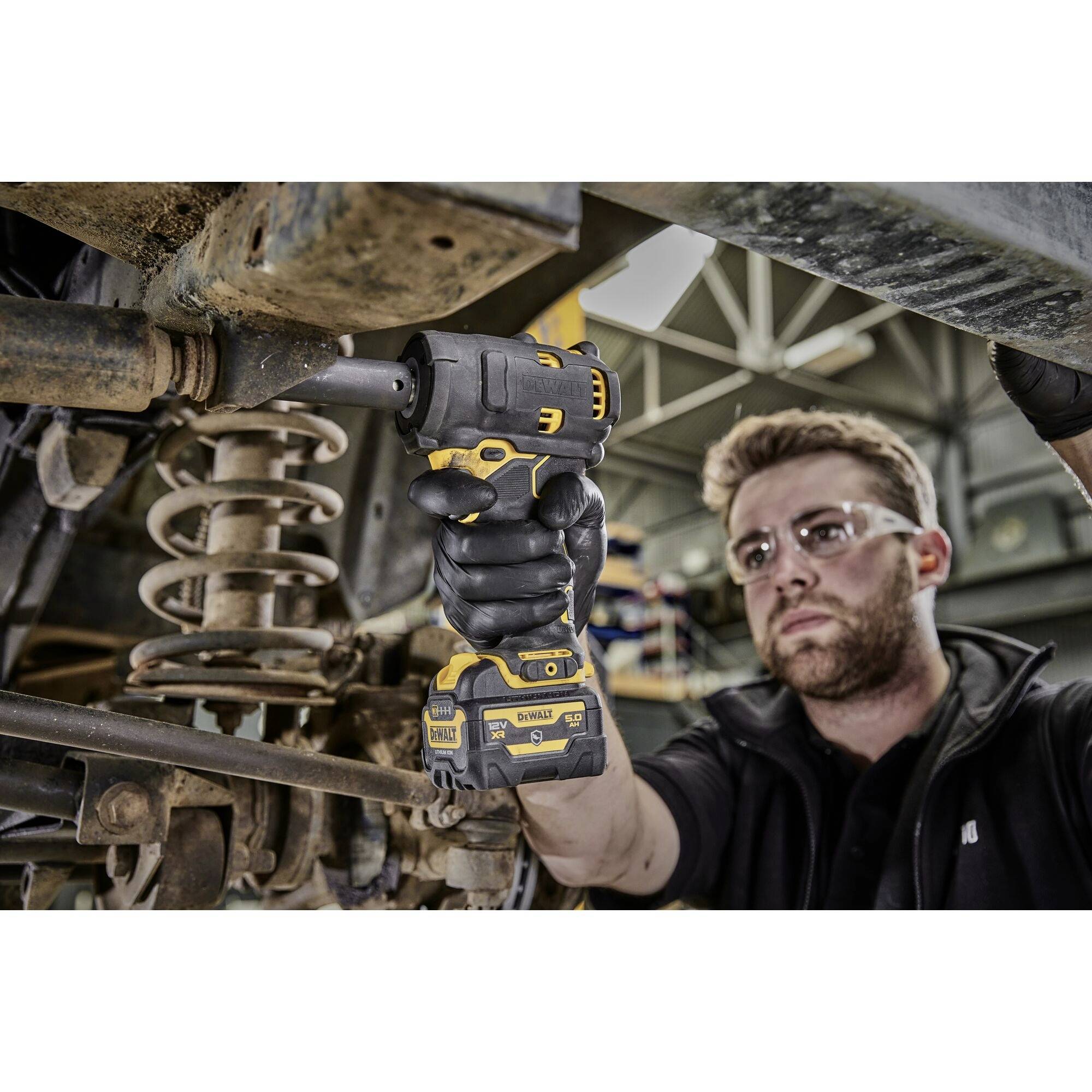 Osoba w okularach ochronnych używa akumulatorowego wkrętarki podczas pracy przy zawieszeniu pojazdu. W tle widoczne jest wnętrze warsztatu.