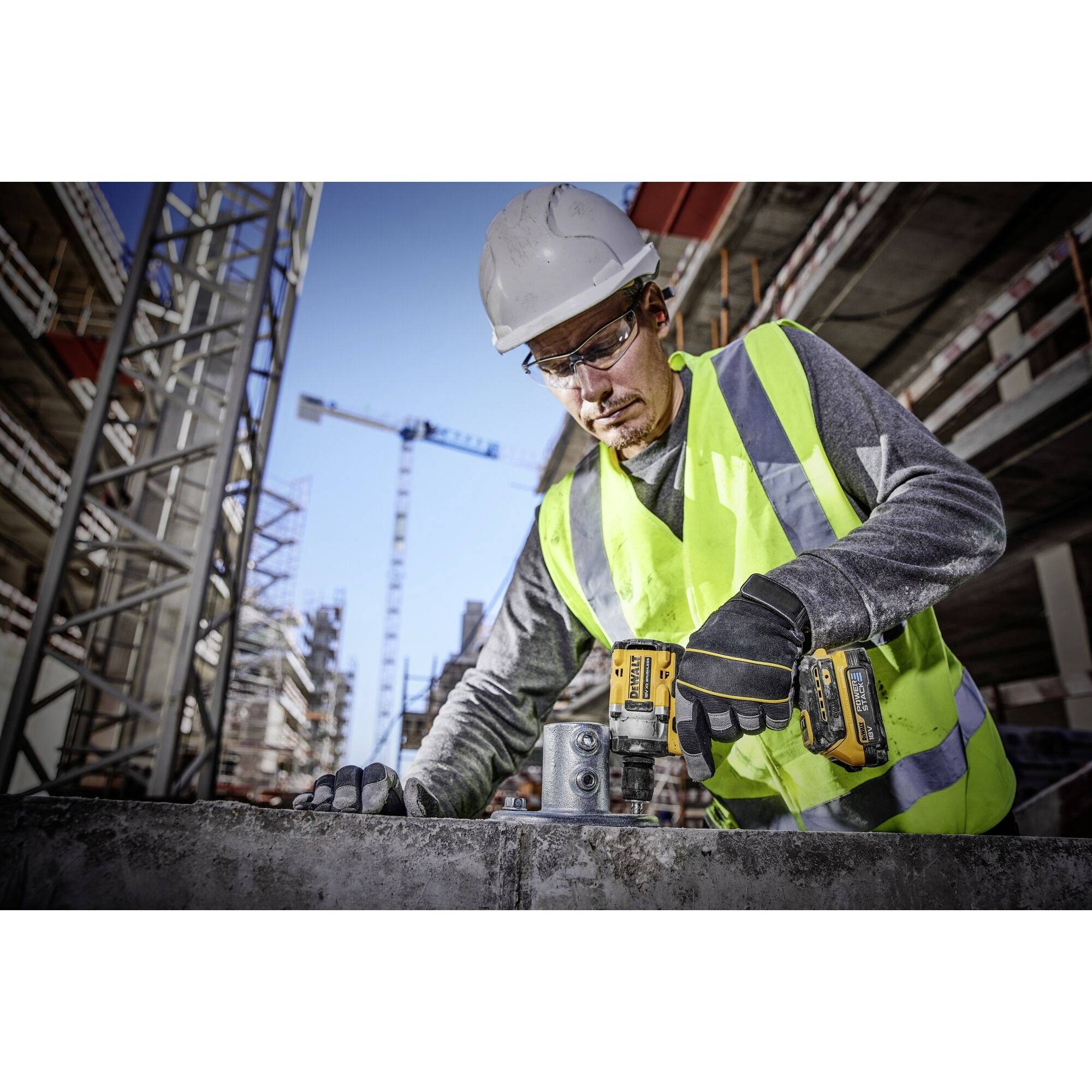 Robotnik budowlany w odzieży ochronnej używa wiertarki na placu budowy. W tle widoczne są dźwigi budowlane i częściowo ukończone budynki.