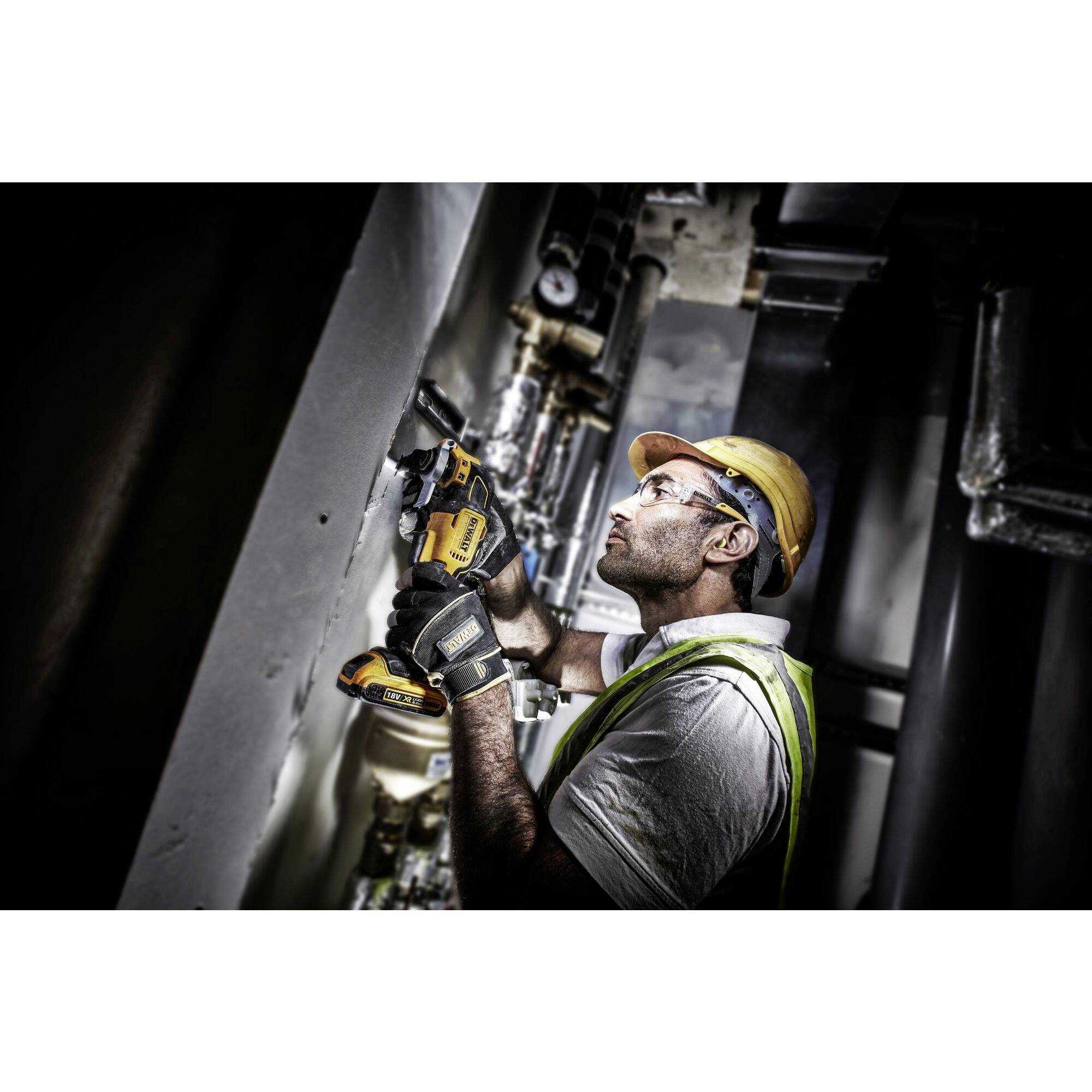 Robotnik budowlany w ochronnych okularach i kasku wywiera nacisk na elektryczną wiertarkę, wykonując otwór w ścianie. Ma na sobie kamizelkę odblaskową, dbając o zachowanie zasad bezpieczeństwa pracy.