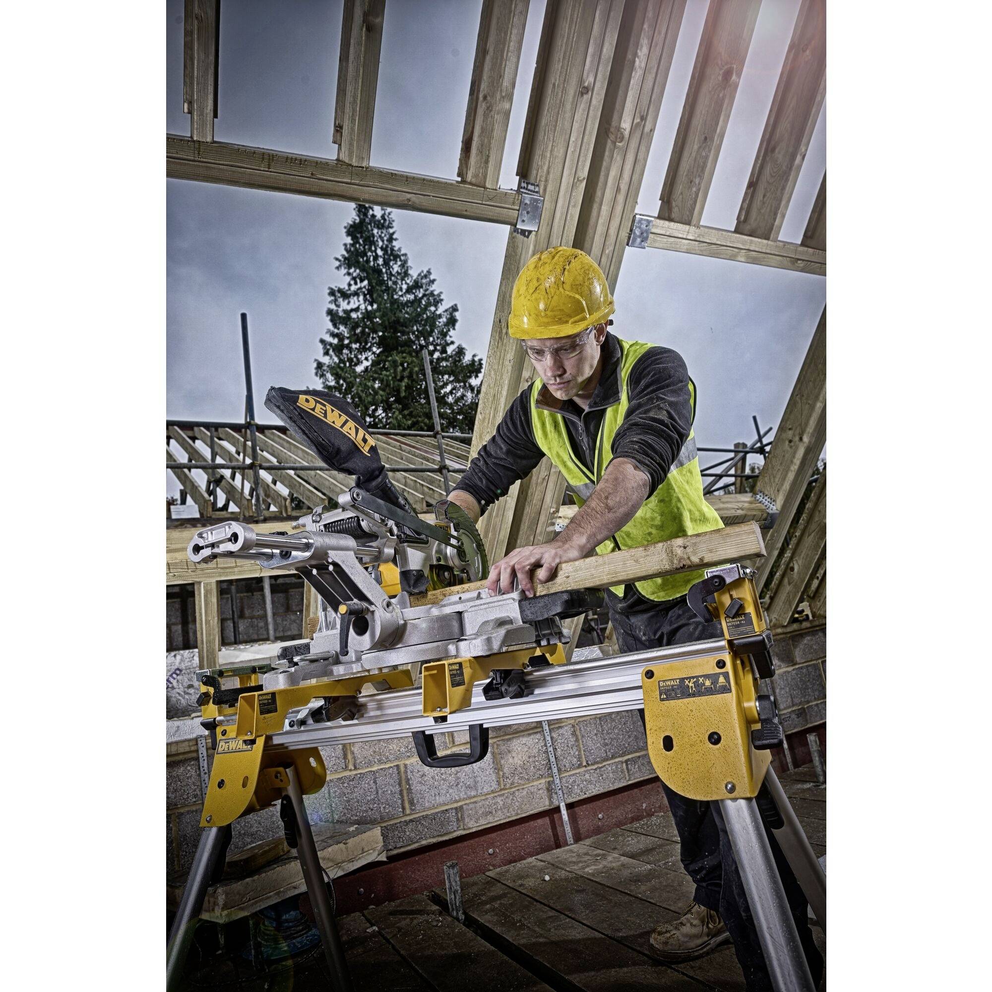 Robotnik w żółtym kasku budowlanym i kamizelce bezpieczeństwa przecina drewno piłą na placu budowy. W tle częściowo zbudowane rusztowanie dachowe.