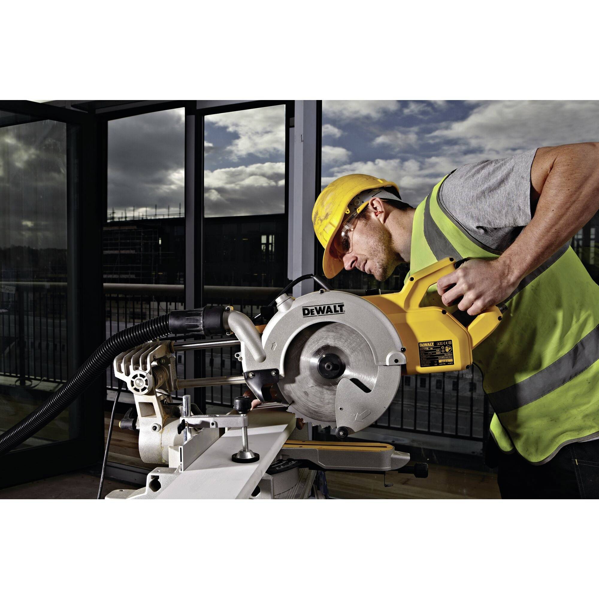 Robotnik budowlany w kasku ochronnym i okularach ochronnych obsługuje elektryczną piłę ukośnicową na placu budowy, rzeżąc drewno.