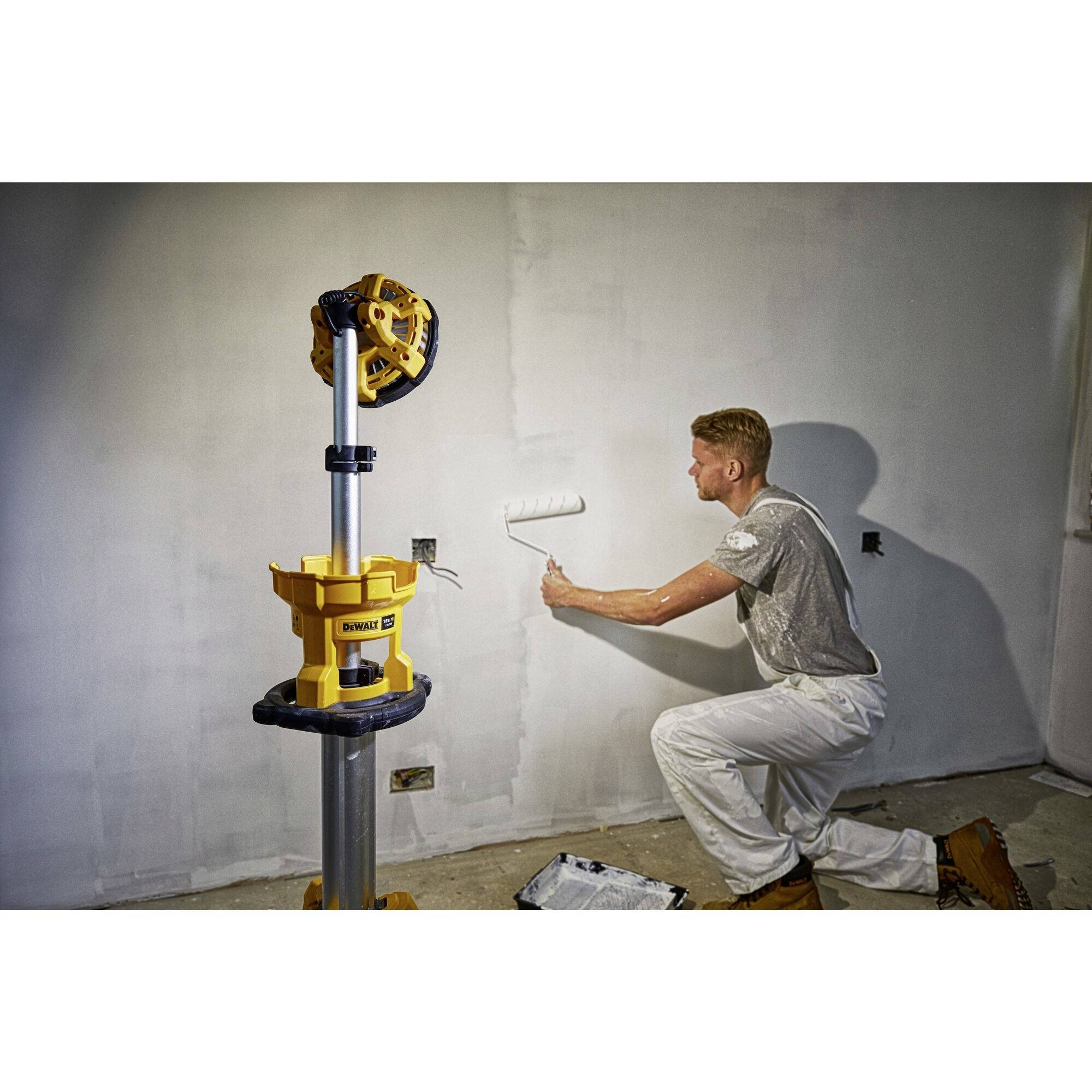 Mężczyzna klęczy i wałkiem malarskim maluje ścianę na biało. Obok niego na statywie stoi żółta lampa budowlana.