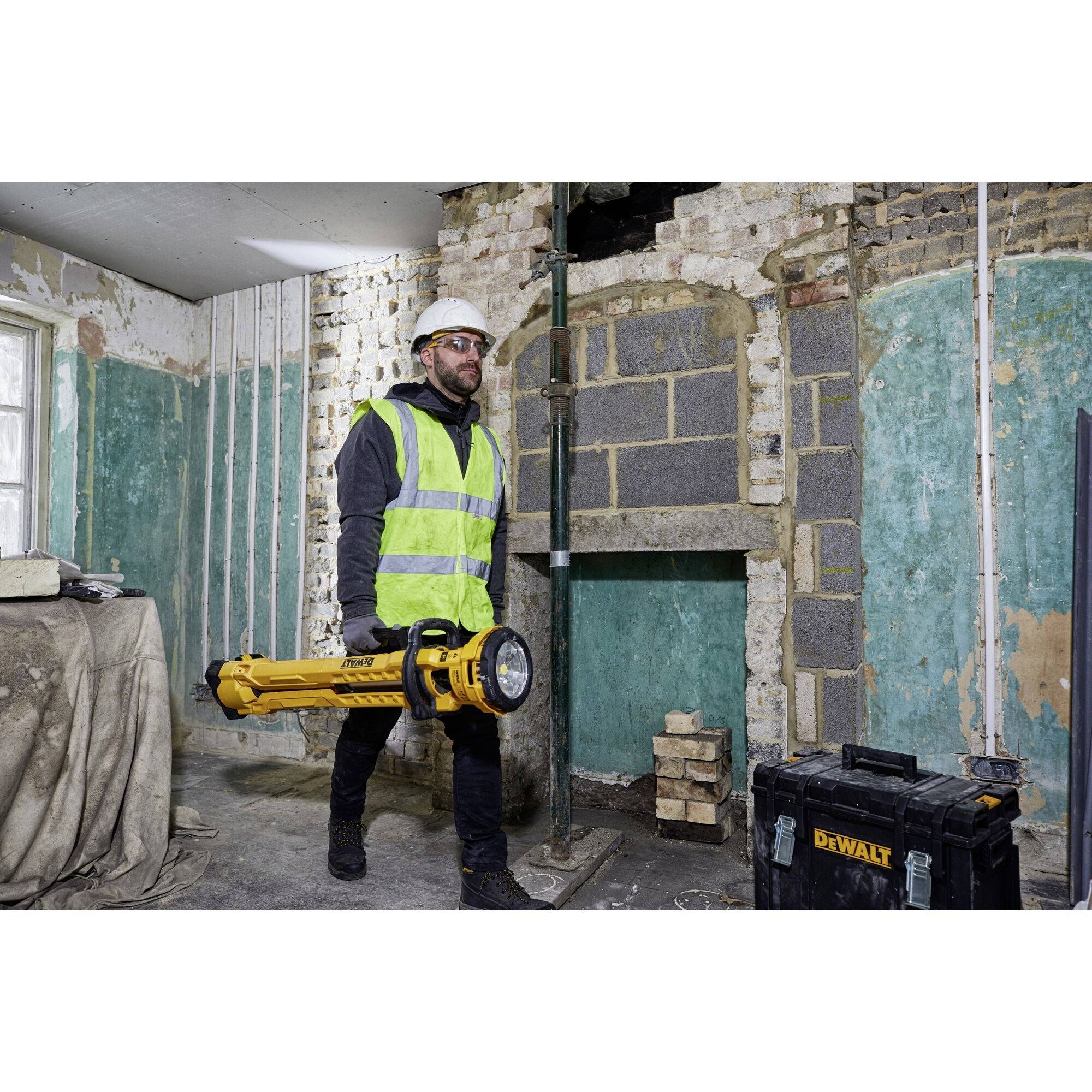 Osoba w sprzęcie ochronnym niesie żółte narzędzie w odnowionym pomieszczeniu z odkrytymi ścianami i narzędziami budowlanymi.