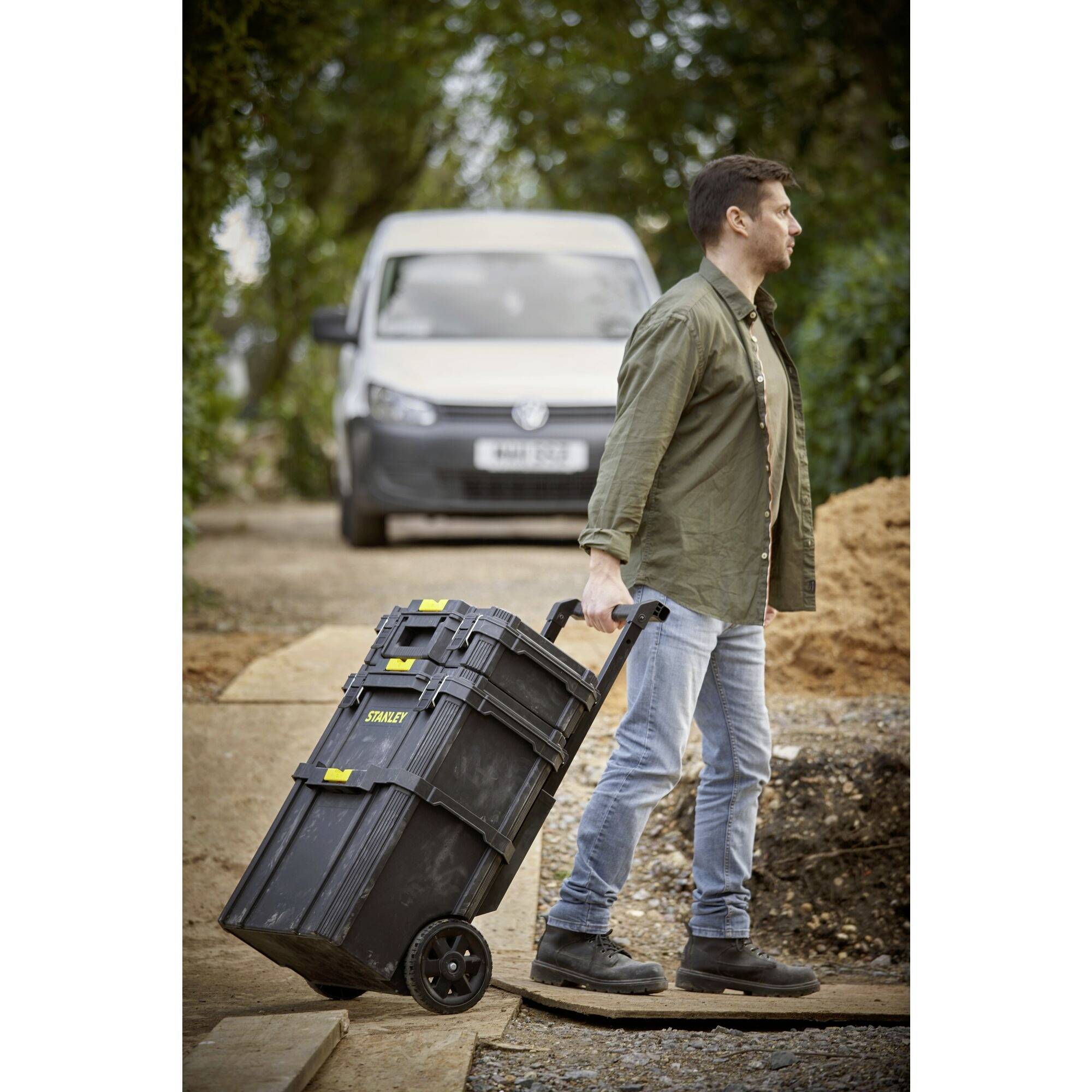 Mężczyzna ciągnie czarną skrzynkę z narzędziami na kółkach po drewnianej pochylni, w tle stoi biały van na budowie.