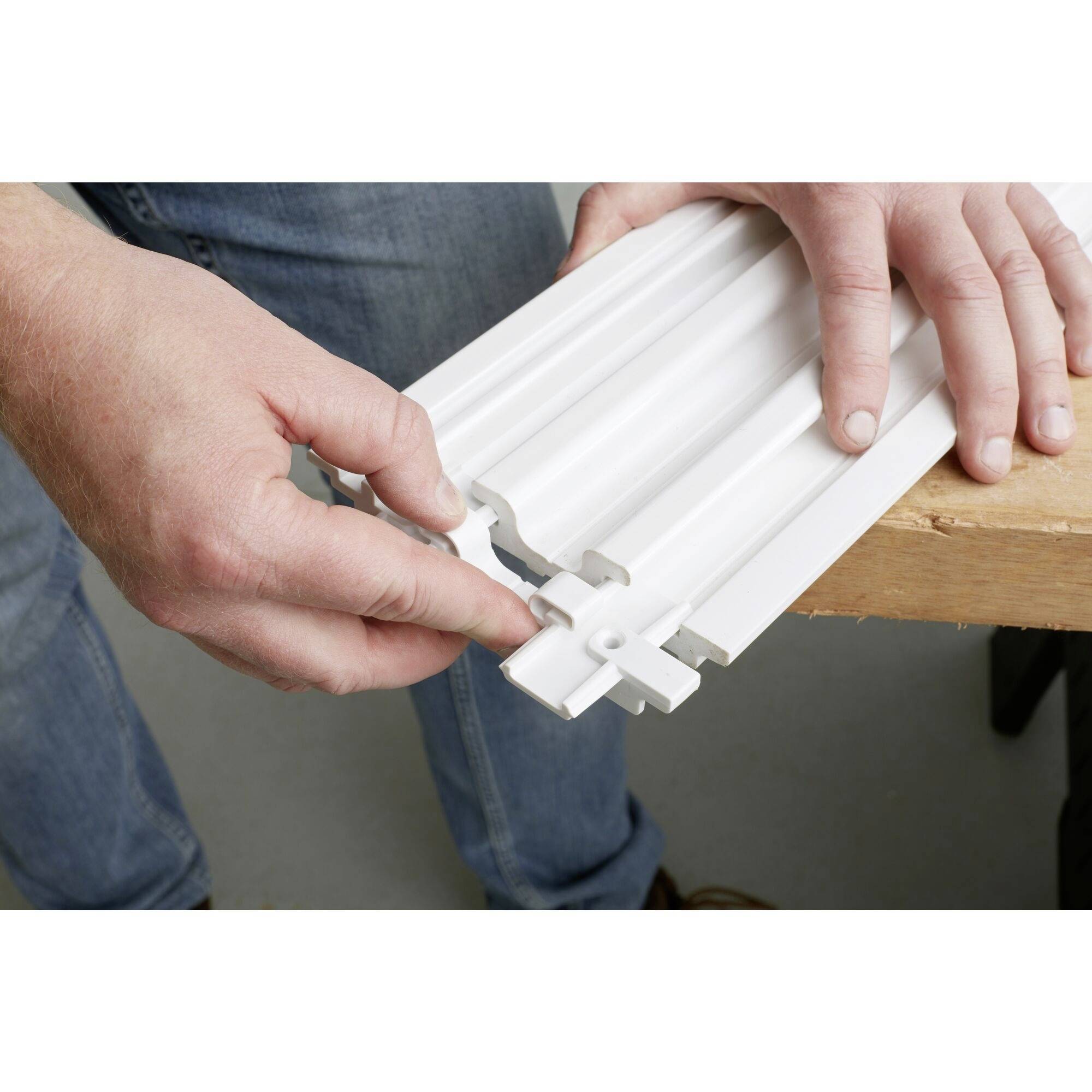 Osoba montuje białą płytę sufitową, łącząc elementy nad stołem. Ręce wskazują na złącza.