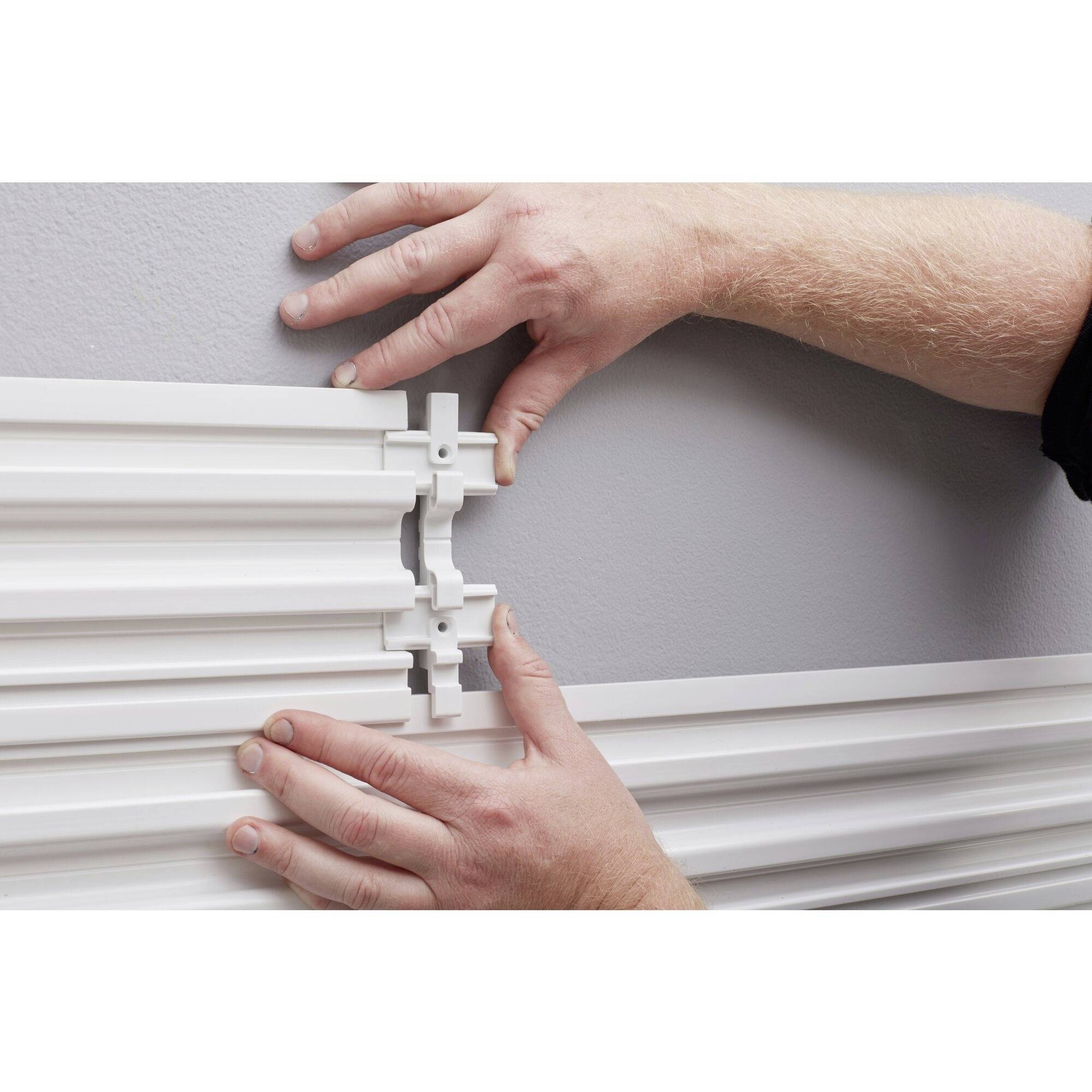 Osoba montuje białą plastikową szynę na szarej ścianie, łącząc dwie szyny za pomocą elementu łączącego.