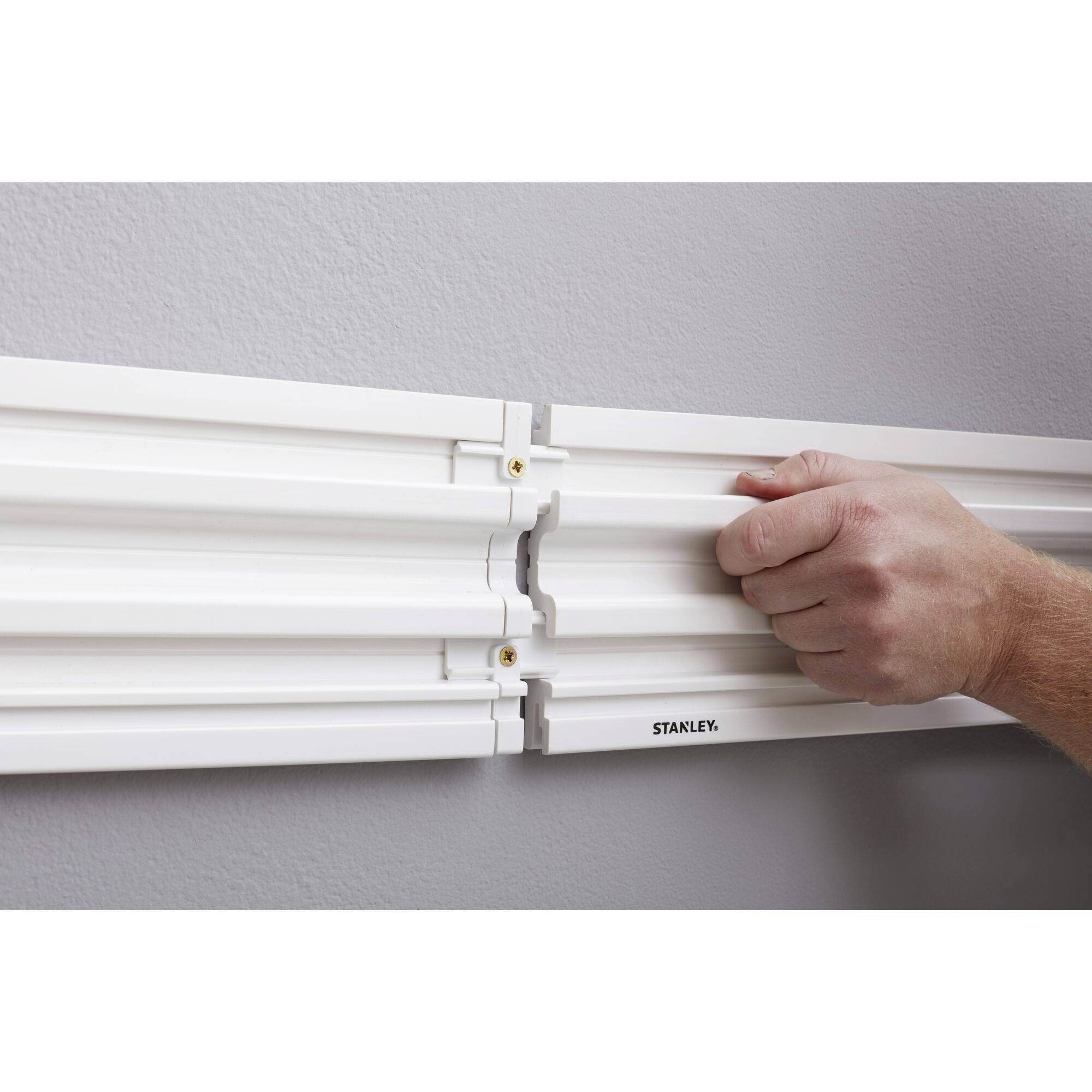 Dłoń przesuwa dwie białe szyny na szarej ścianie, które są łączone. Na jednej szynie widnieje napis 'STANLEY'.