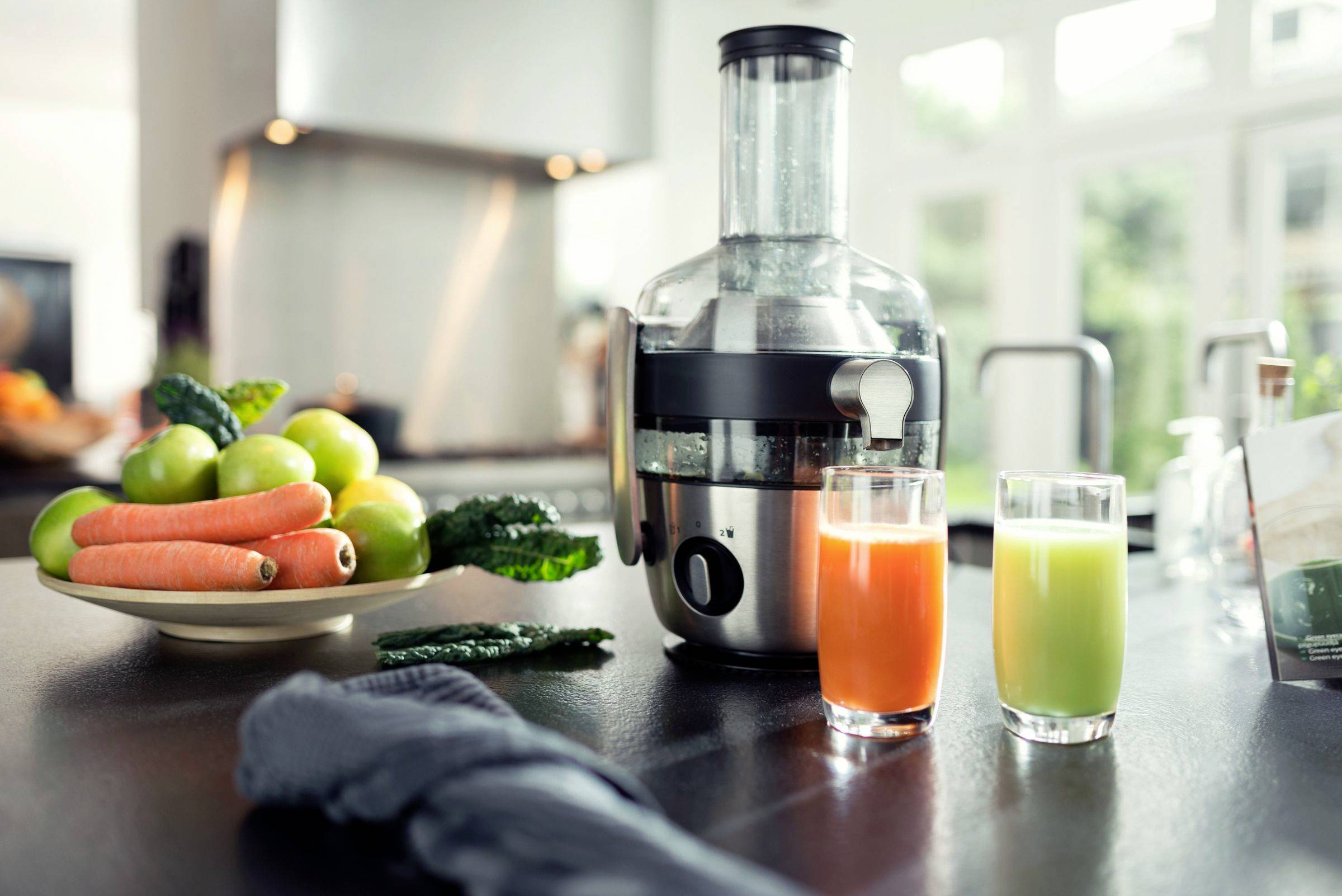 Sokowirówka stoi na blacie kuchennym obok dwóch szklanek soku, jednej pomarańczowej i jednej zielonej, oraz talerza z owocami i warzywami.