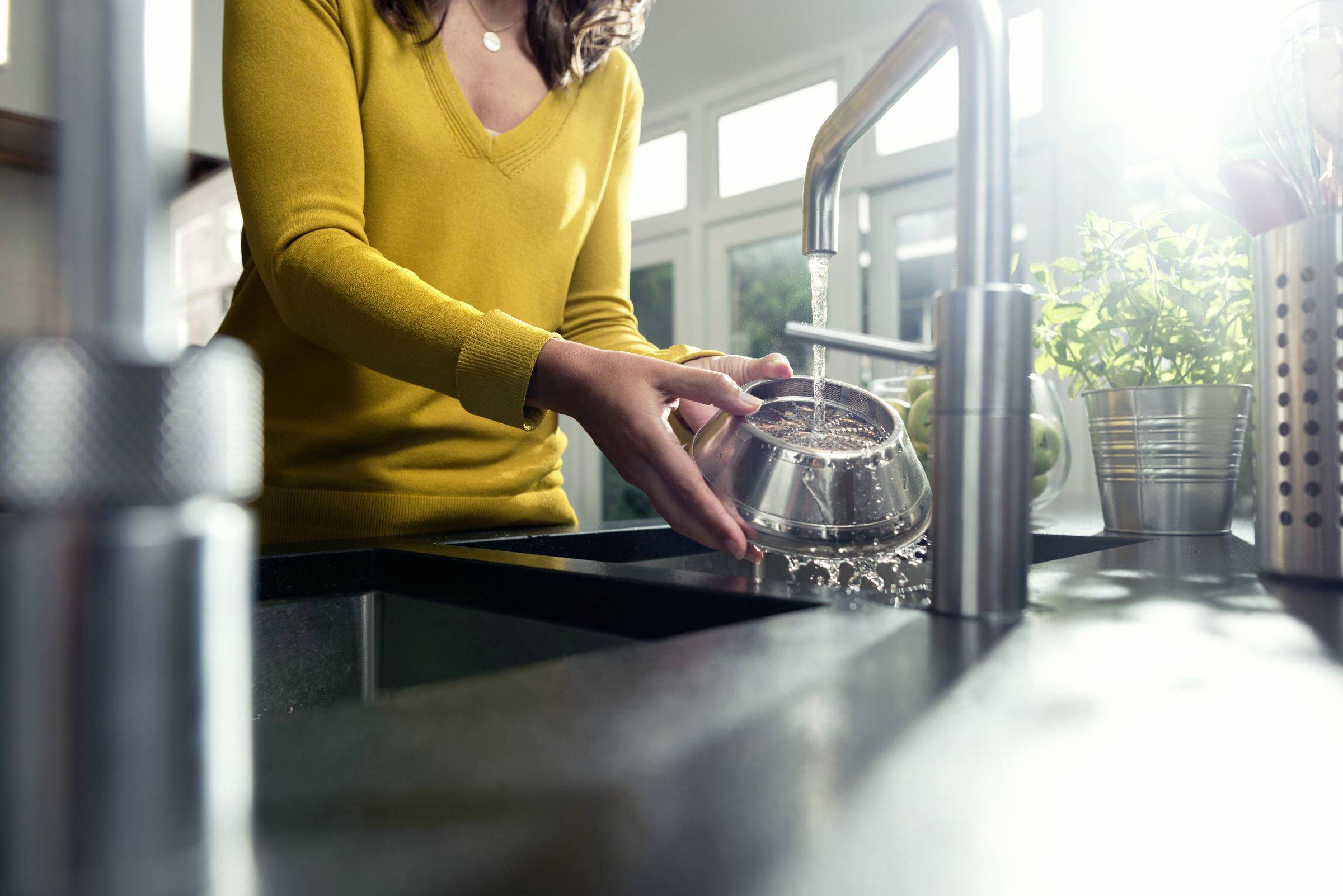 Osoba myje metalową miskę pod bieżącą wodą w kuchni. W tle rośliny, przez okno wpada słońce.