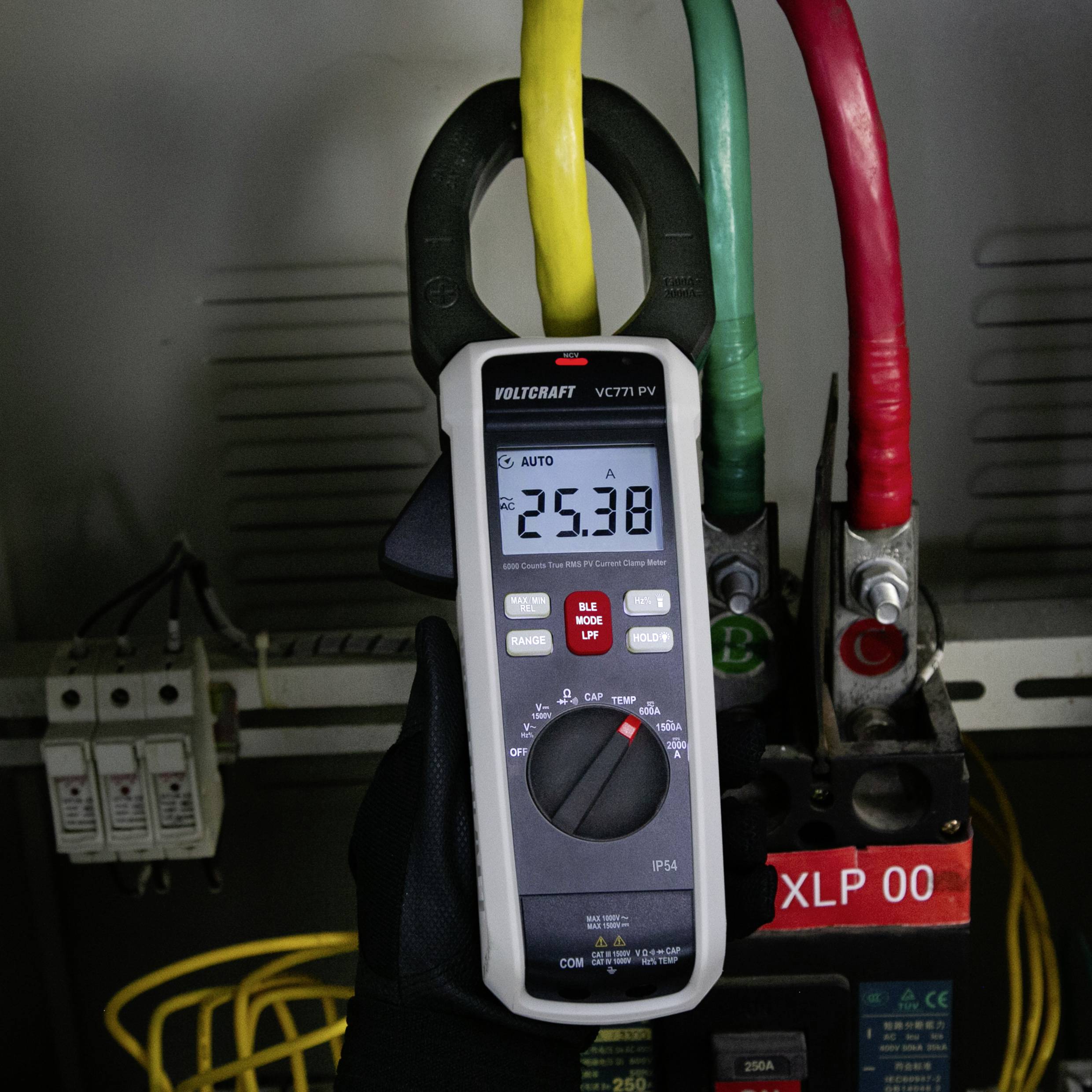 Multimetr sprawdza napięcie, wyświetlając '25,38' na tablicy elektrycznej z podłączonymi przewodami żółtym, zielonym i czerwonym.