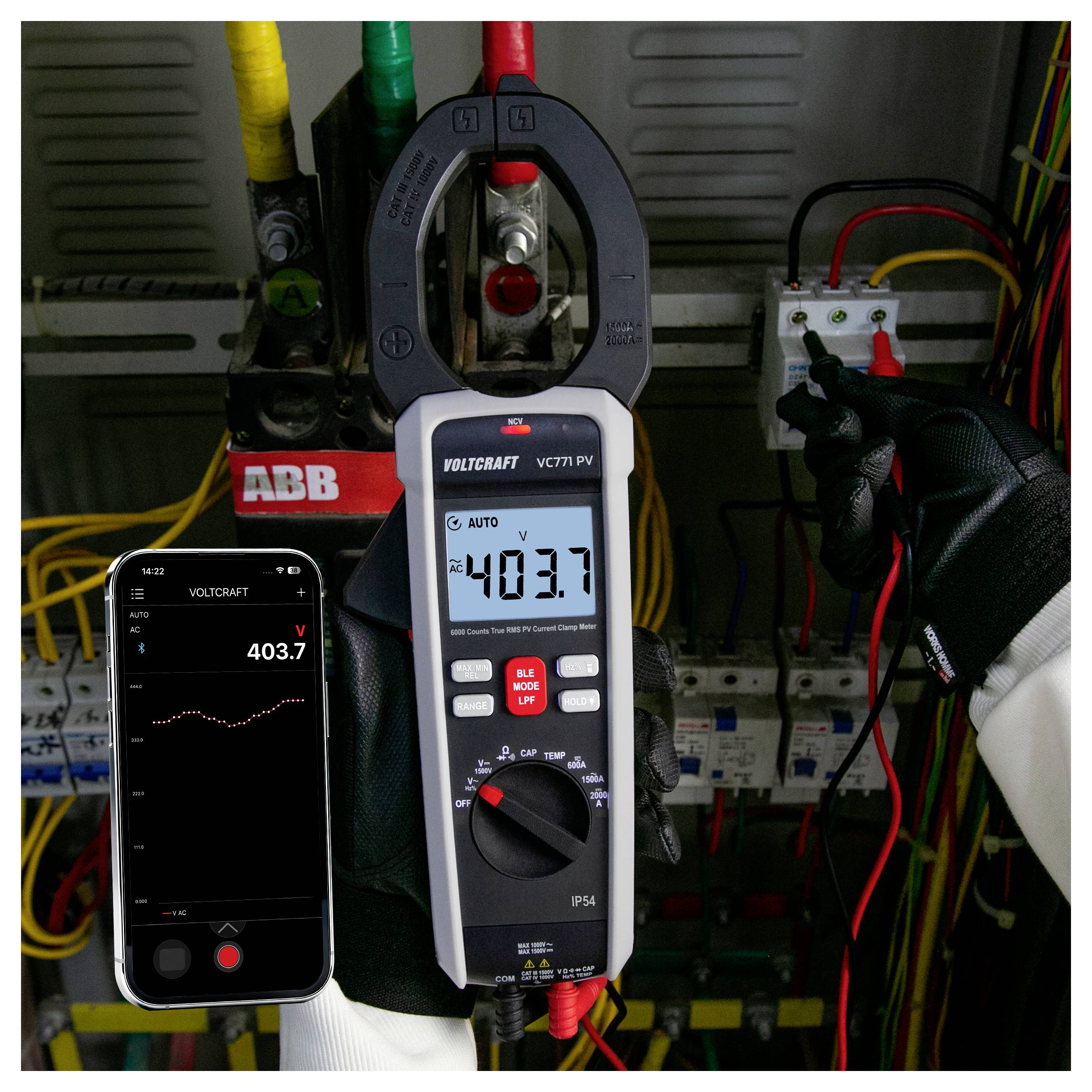 Technik mierzy napięcie elektryczne w rozdzielni za pomocą miernika cęgowego i smartfona, oba urządzenia wyświetlają około 403,7 V.