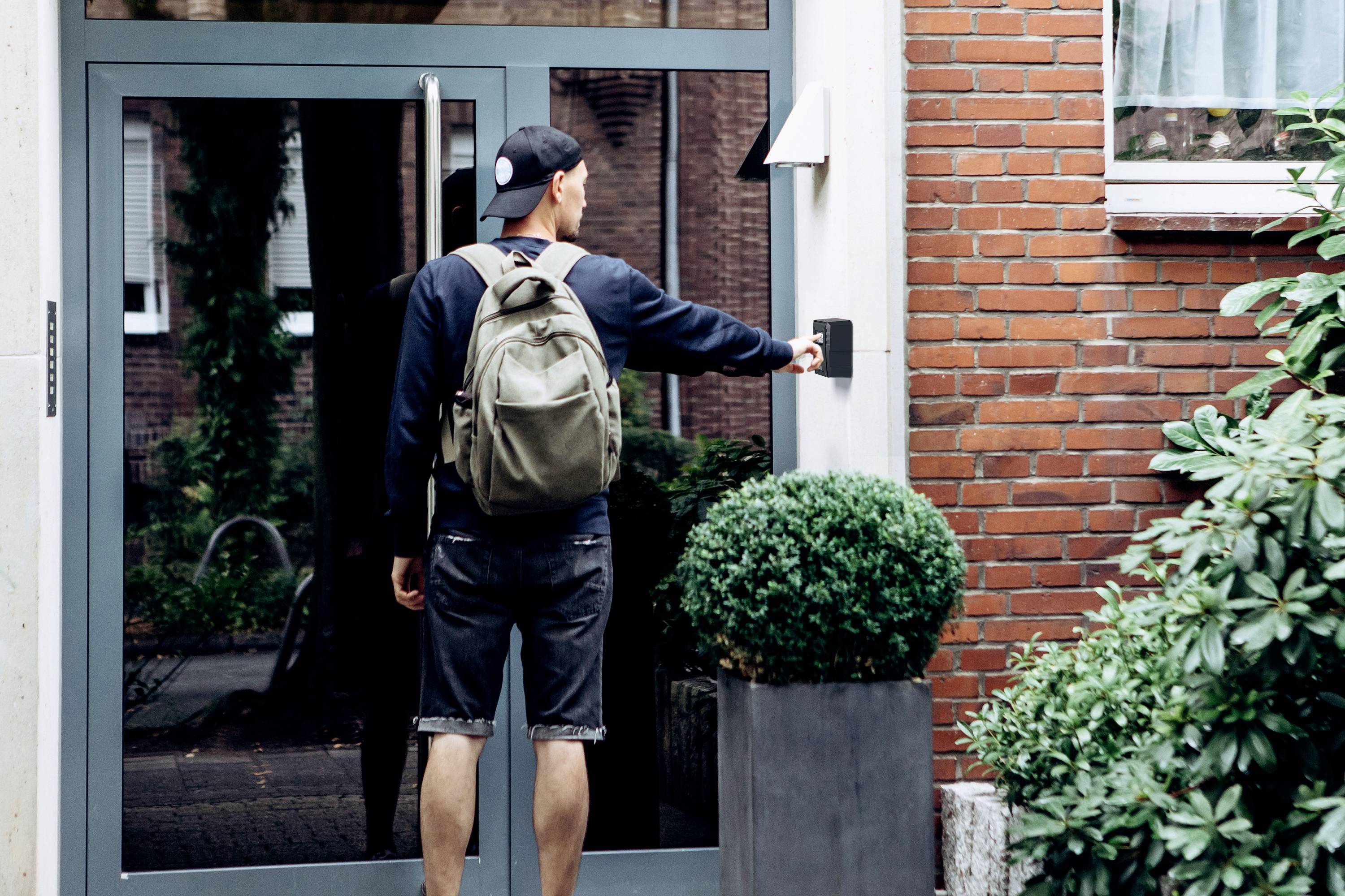 Osoba z plecakiem dzwoni do drzwi wejściowych obok ceglastego muru. Na pierwszym planie widoczne są doniczkowe rośliny.