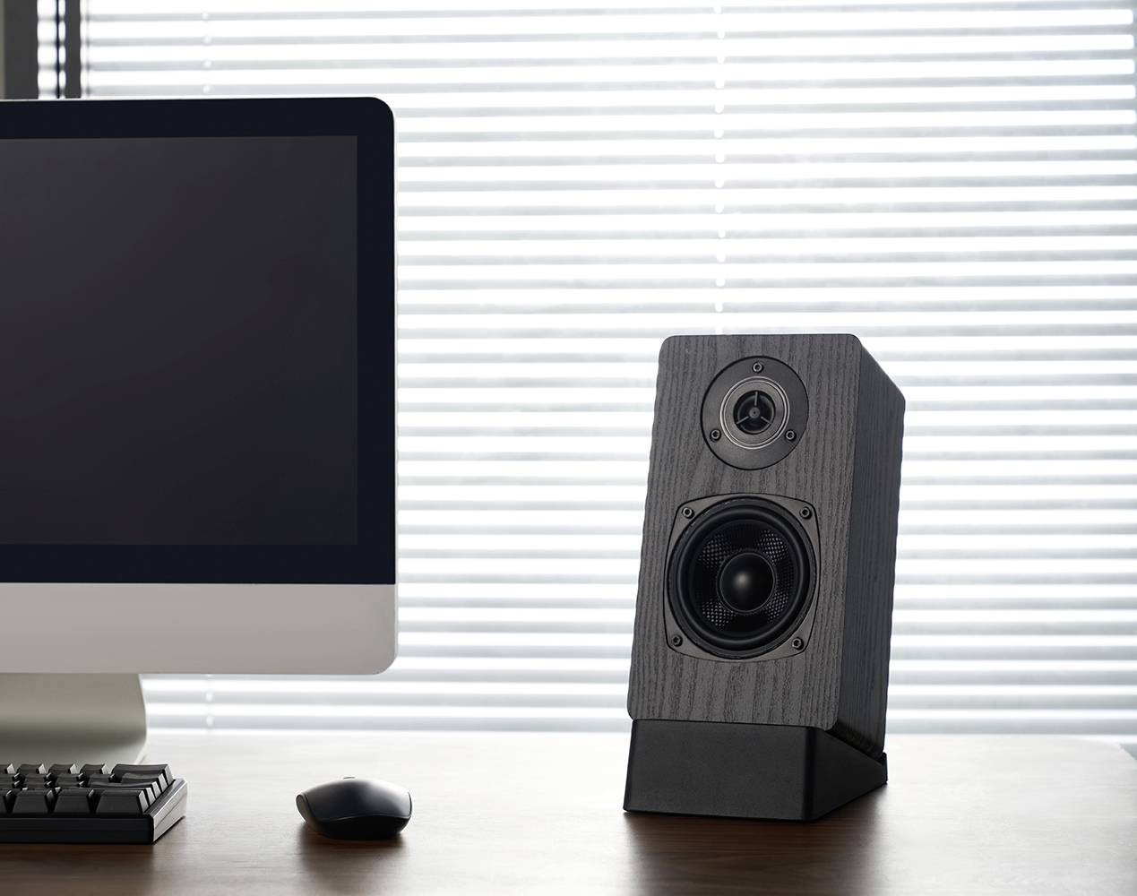 Na biurku przed oknem z żaluzjami stoi ekran komputerowy i głośnik. Scena wygląda nowocześnie i minimalistycznie.