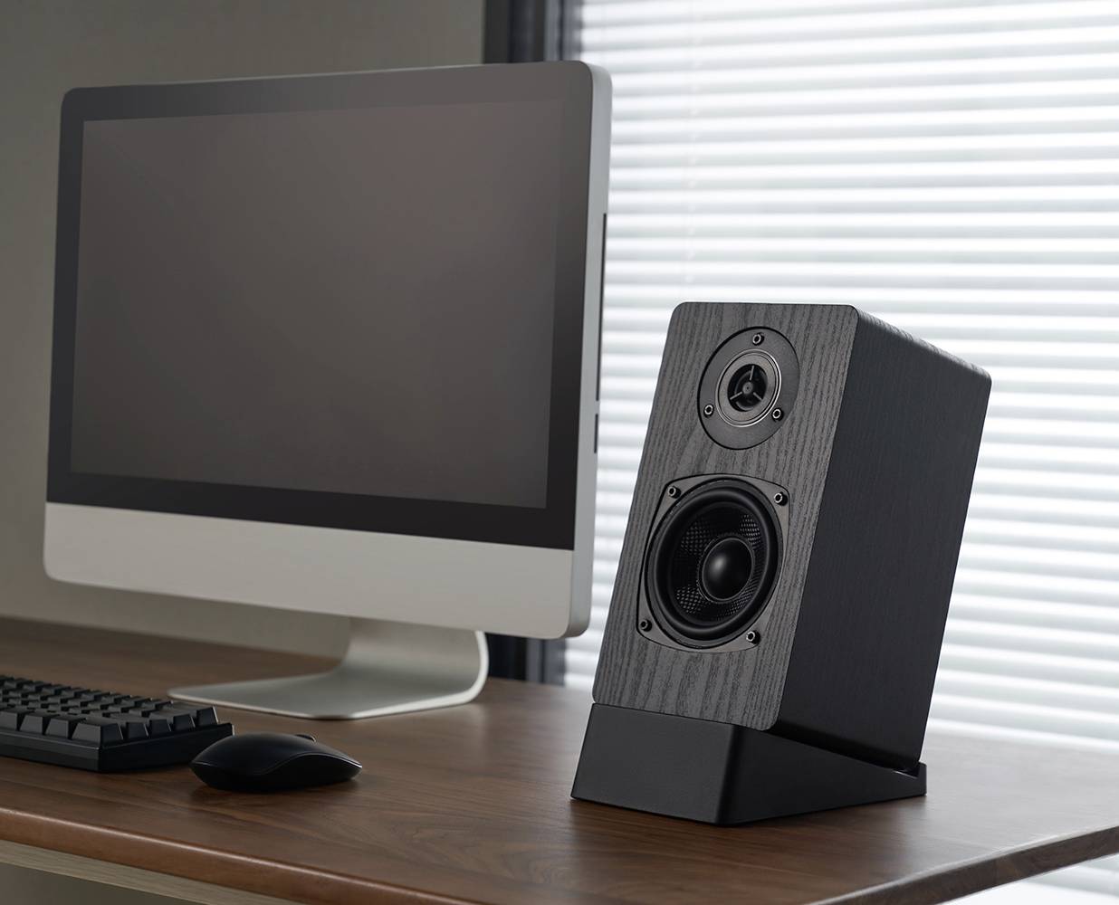 Komputer z monitorem i klawiaturą na drewnianym biurku. Po prawej stronie stoi czarny głośnik. W tle żaluzje.