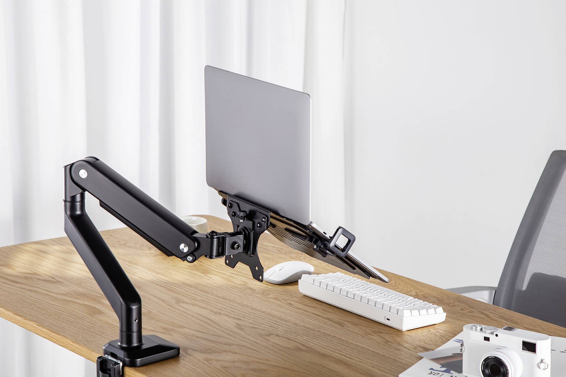 Regulowany na wysokość, czarny uchwyt monitora trzyma laptopa na drewnianym stole. Obok znajdują się klawiatura i kamera.