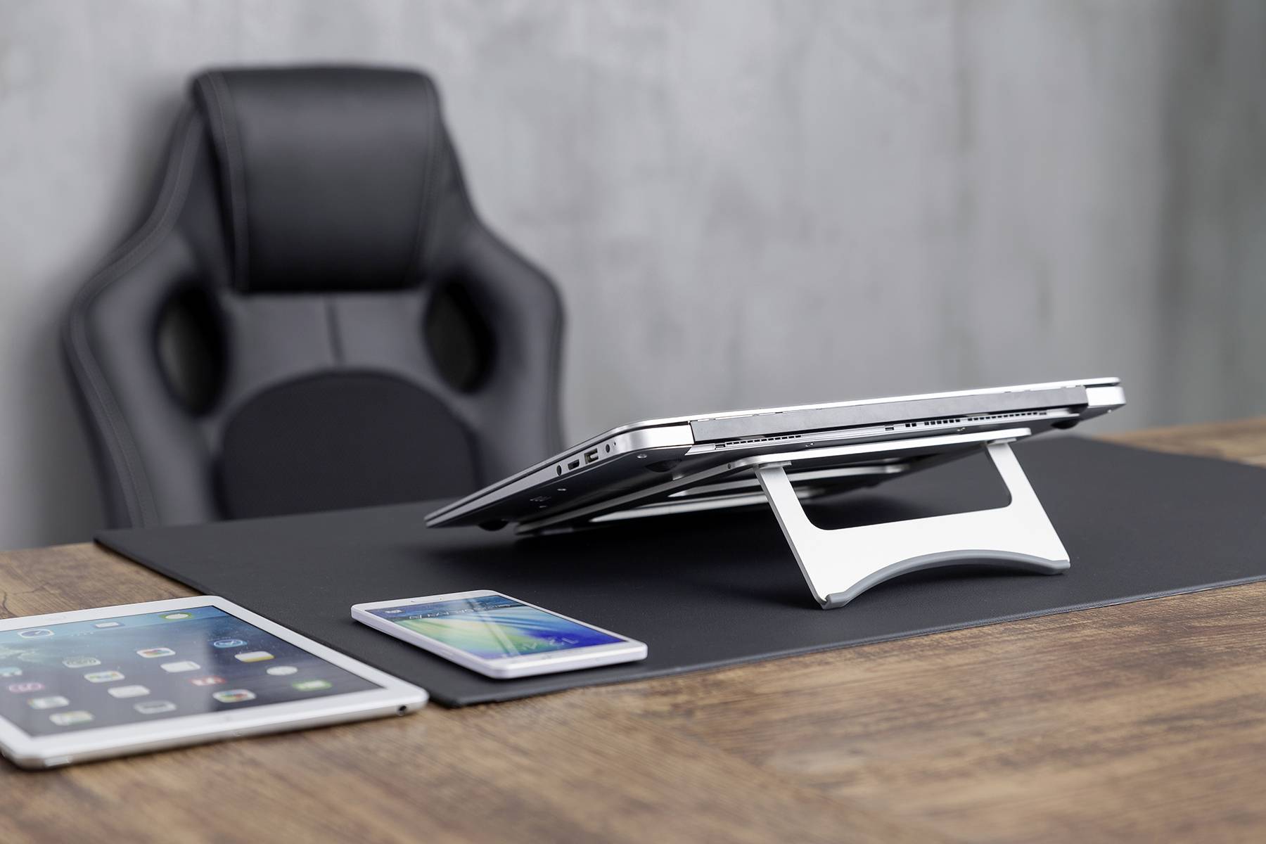 Laptop stoi na białym stojaku na biurku, obok niego tablet i smartfon, w tle czarny fotel biurowy.