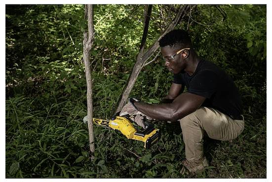Osoba używająca piły do cięcia gałęzi w lesie, nosząca okulary ochronne i rękawice, otoczona zieloną roślinnością.