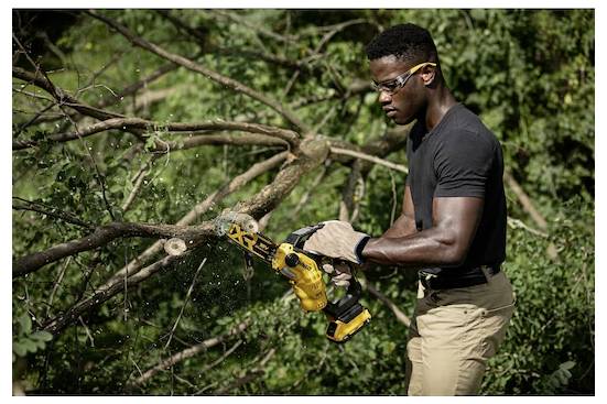 Osoba w okularach ochronnych i rękawicach używa żółtej elektrycznej piły do cięcia gałęzi w zalesionym terenie.