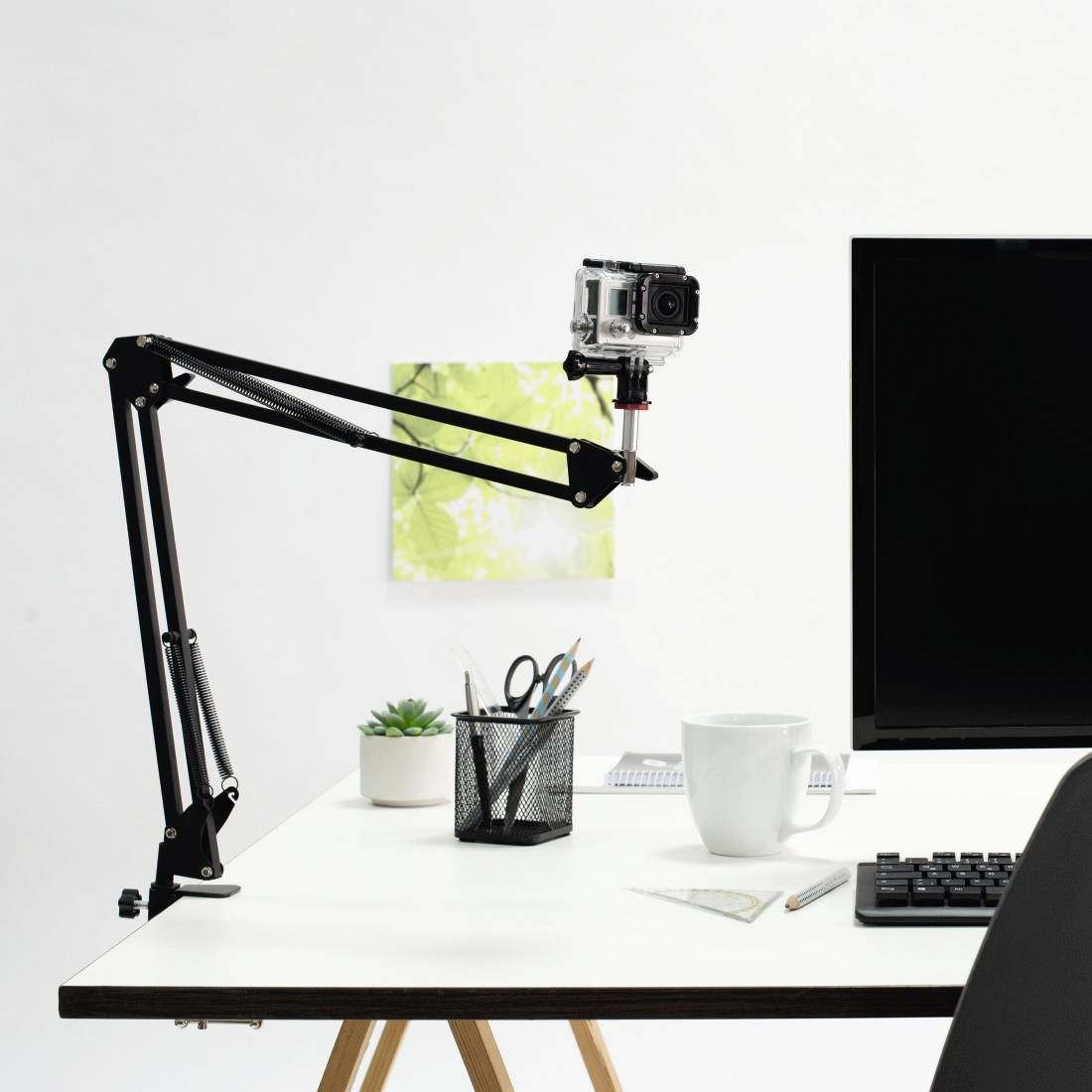 Biurko z białą powierzchnią, na którym znajduje się ramię mikrofonowe z kamerą, rośliny, pojemnik na przybory biurowe, kubek i klawiatura.