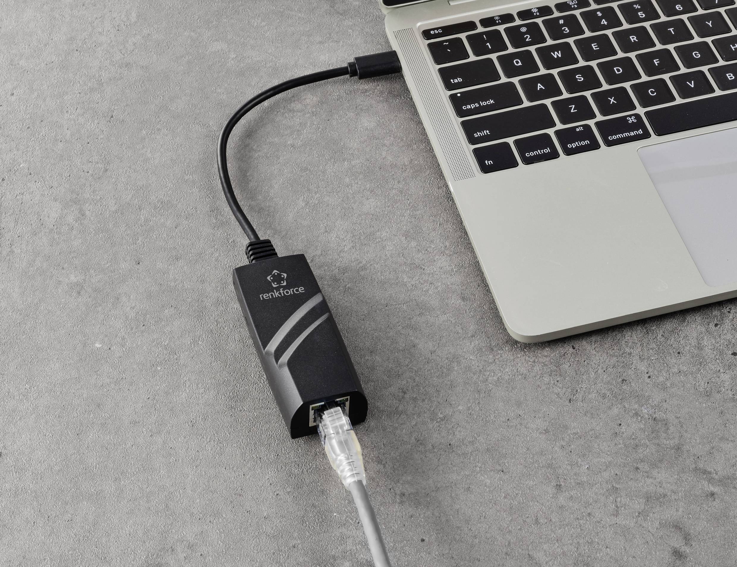 Laptop jest podłączony kablem Ethernet przez adapter USB, który leży na szarym stole.