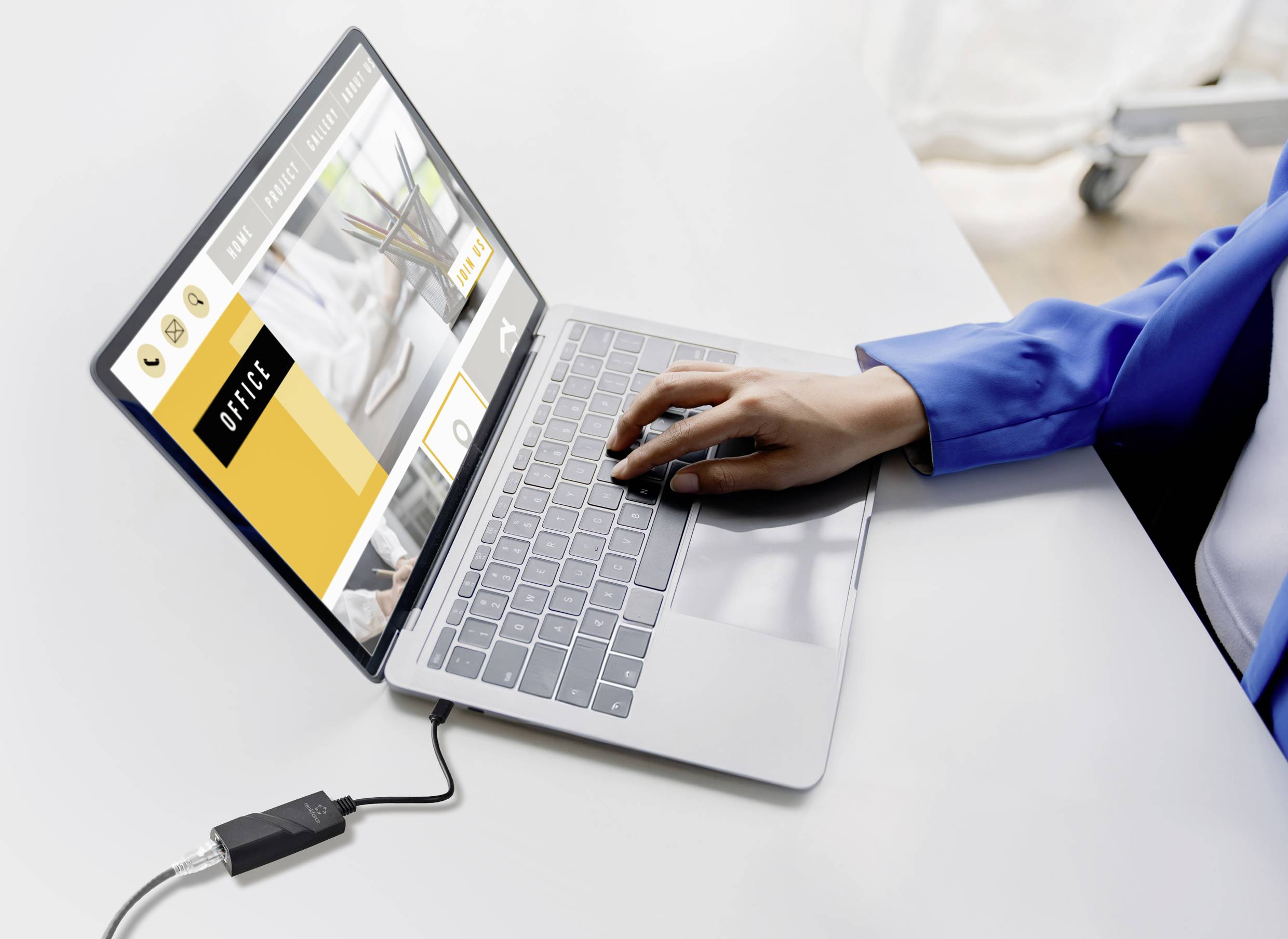 Osoba w niebieskiej marynarce pisze na laptopie z otwartą stroną internetową. Laptop jest podłączony do adaptera.