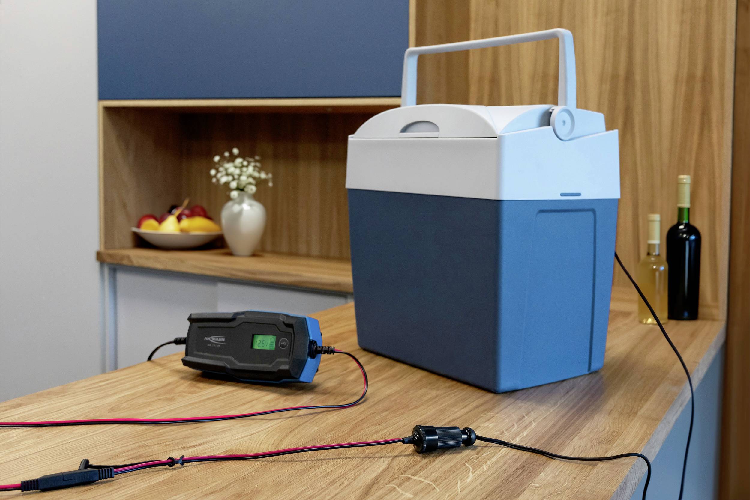 Niebieska lodówka turystyczna z pokrywą stoi na drewnianym blacie, podłączona do ładowarki. W tle znajduje się wazon z kwiatami i owocami.