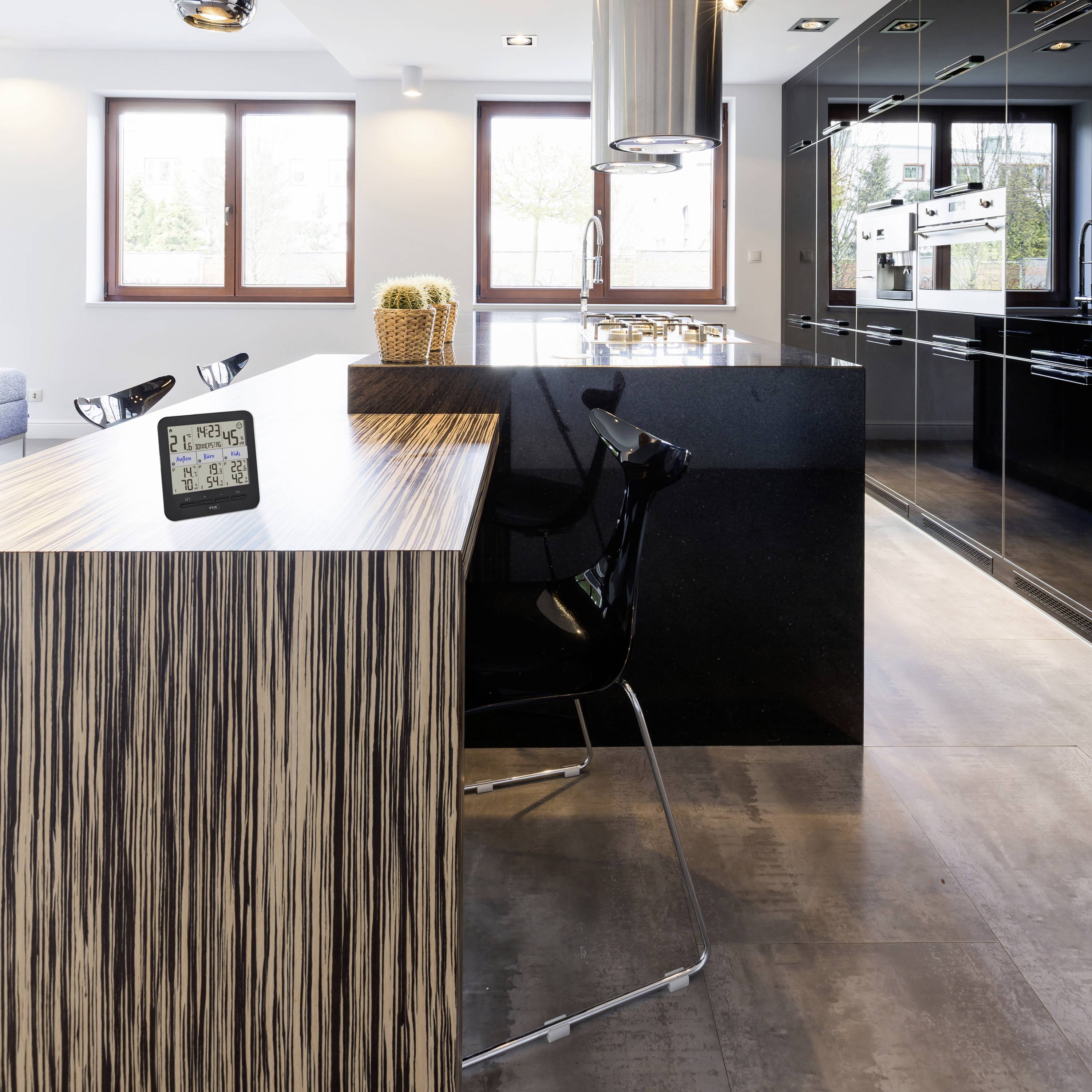 Nowoczesna kuchnia z drewnianym blatem, czarnym hokererem, cyfrowym zegarem i doniczkową rośliną. Duże okna, jasne wnętrze.