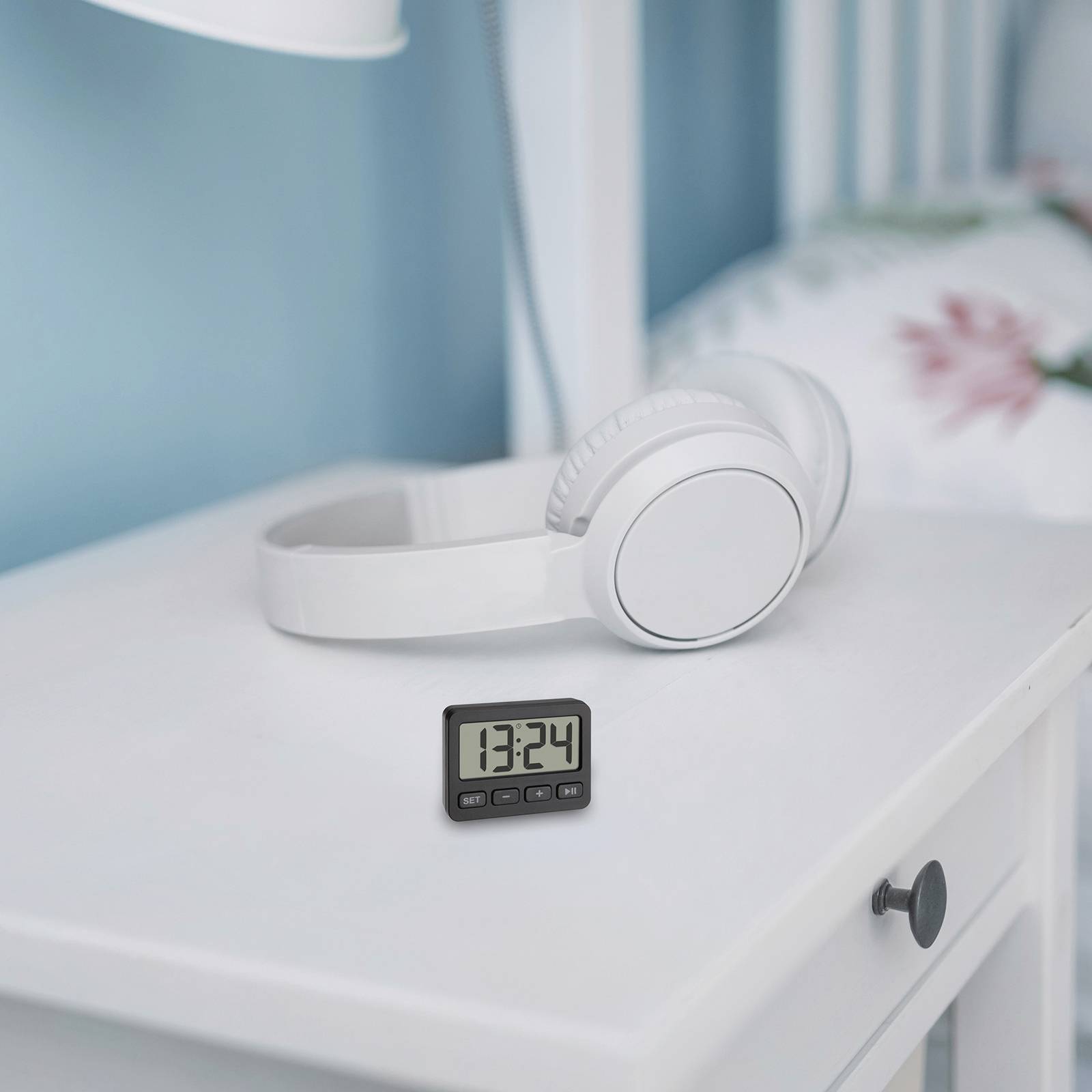 Cyfrowy budzik wskazuje godzinę 13:24 na białym stoliku nocnym. Obok leżą białe słuchawki. W tle widać łóżko.