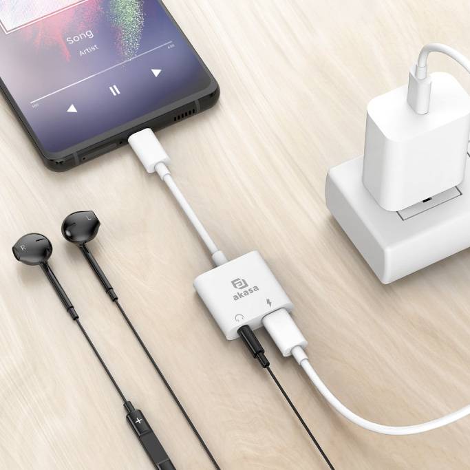 Smartfon z włączoną muzyką, podłączony do stacji ładującej. Obok słuchawki z przewodem. Na jasnej drewnianej powierzchni.