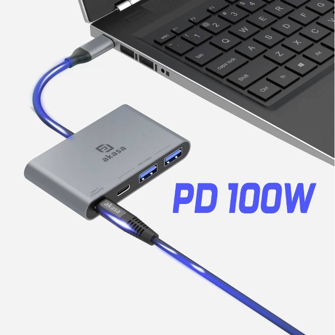 Laptop jest podłączony przez kabel USB-C do szarego adaptera, który posiada dwa porty USB i port ładowania. Tekst: 'PD 100W'.