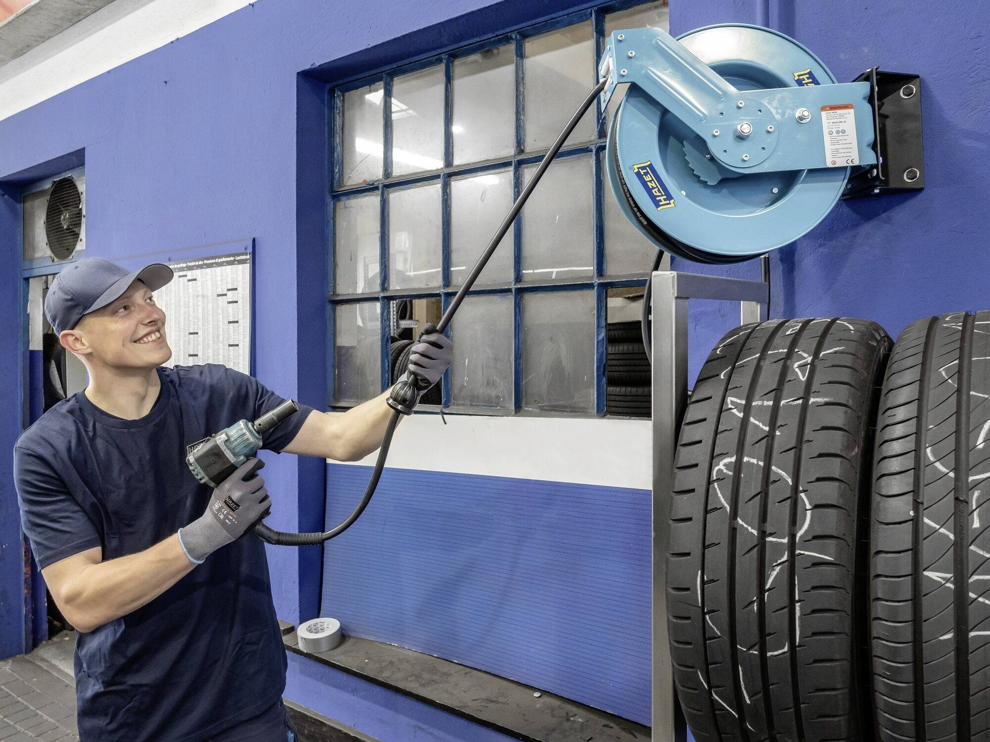 Mężczyzna w stroju roboczym wyciąga kabel z bębna kablowego w warsztacie obok ułożonych stosów opon samochodowych.
