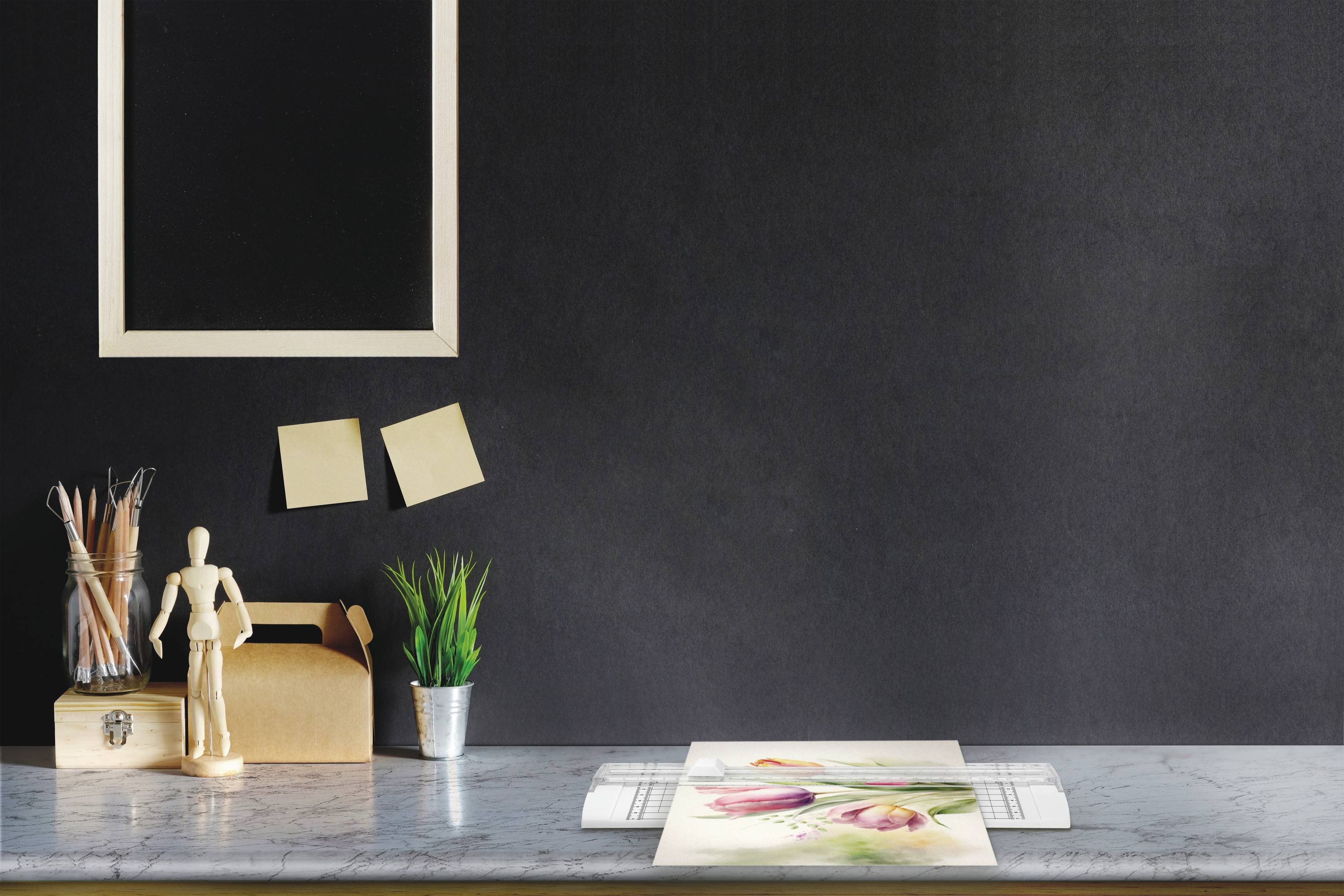 Biurko z przyborami do rysowania, notatnikiem i roślinami. Na ścianie wiszą dwie karteczki samoprzylepne i pusta ramka na zdjęcie.