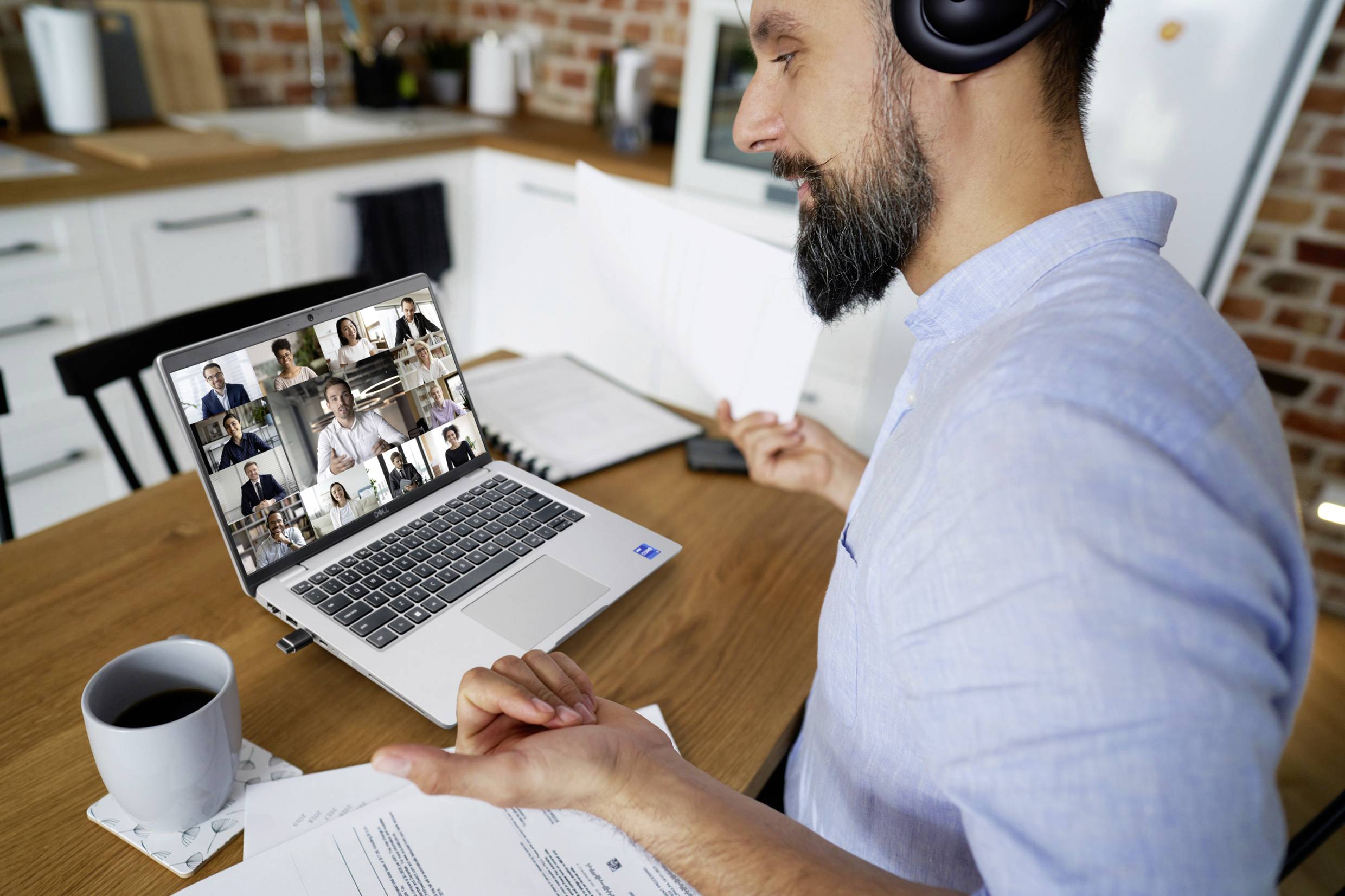 Mężczyzna w słuchawkach siedzi przy stole, trzymając dokumenty, podczas gdy bierze udział w wideokonferencji z wieloma osobami na laptopie.
