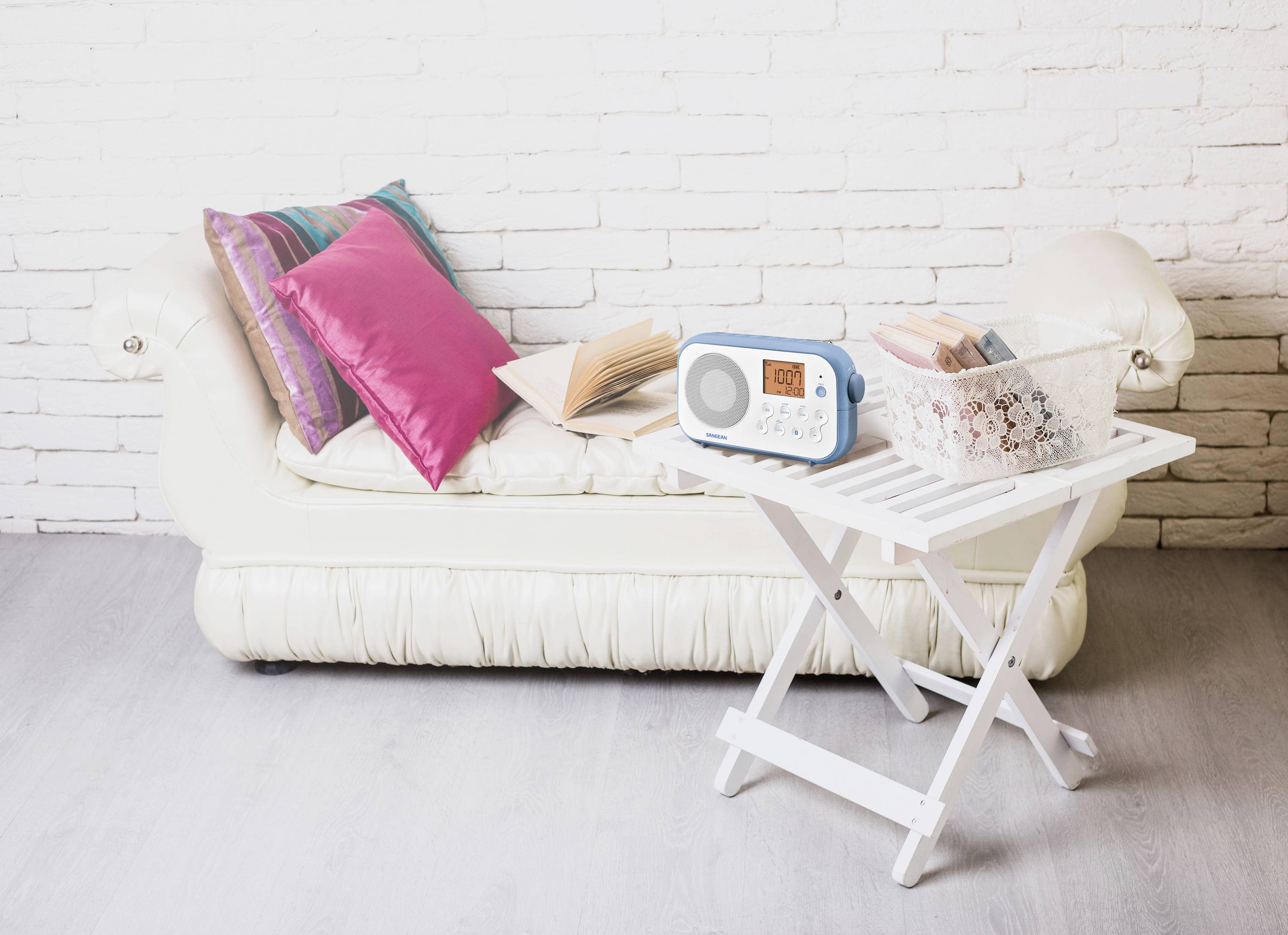 Biała sofa z kolorowymi poduszkami stoi przed białą ścianą z cegły. Na sofie leży otwarta książka. Obok, biały stolik trzyma radio i kosz z wachlarzami.