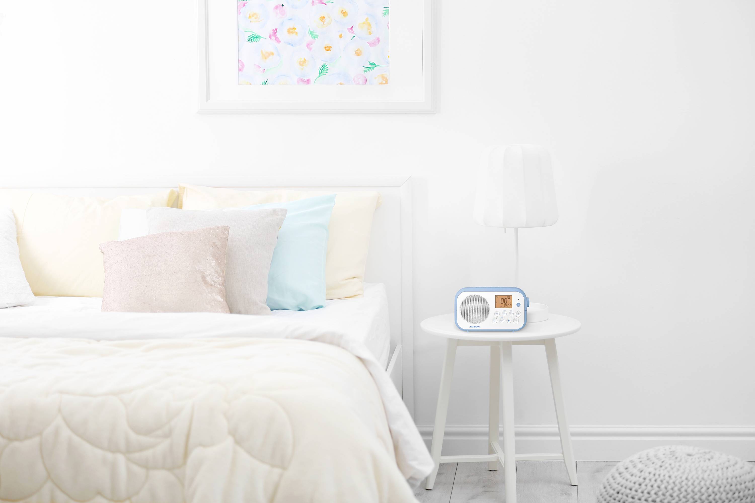 Jasna sypialnia z białym łóżkiem, kolorowymi poduszkami i małym stolikiem, na którym stoi białe radio. Kolorowy obraz na ścianie.