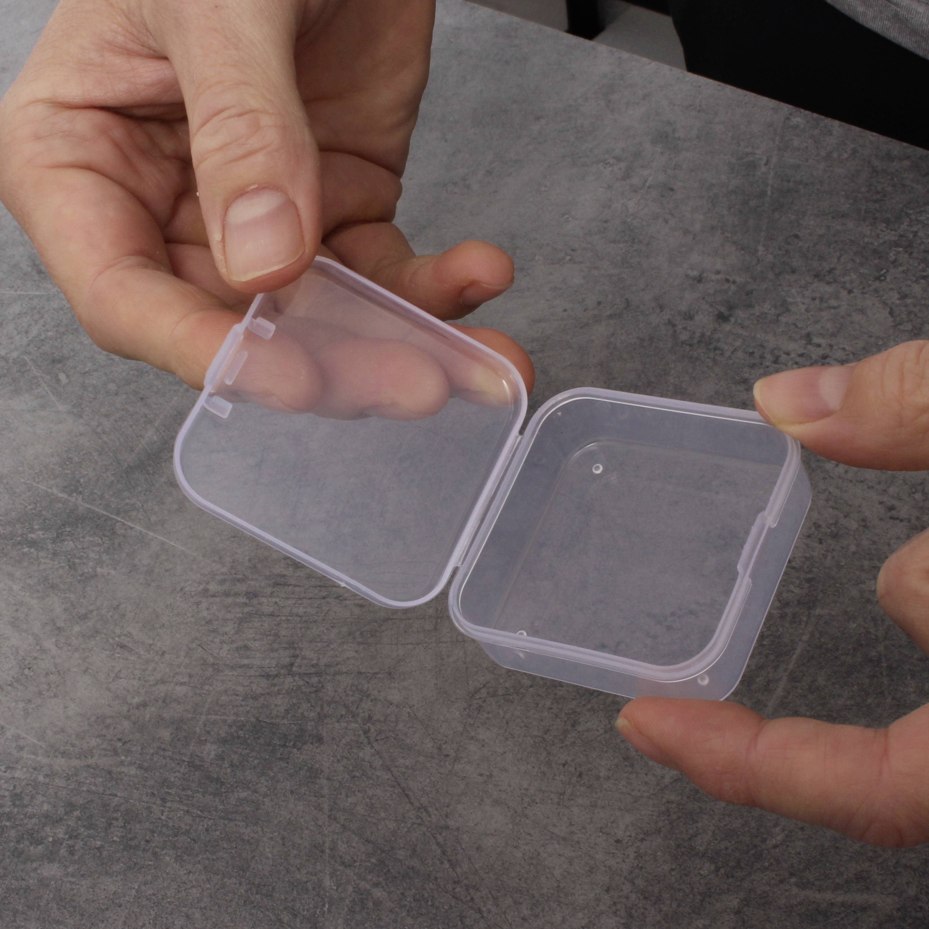 Osoba trzyma otwarte, przezroczyste pudełko z tworzywa sztucznego w ręku, gotowa coś włożyć lub wyjąć, na szarym stole.