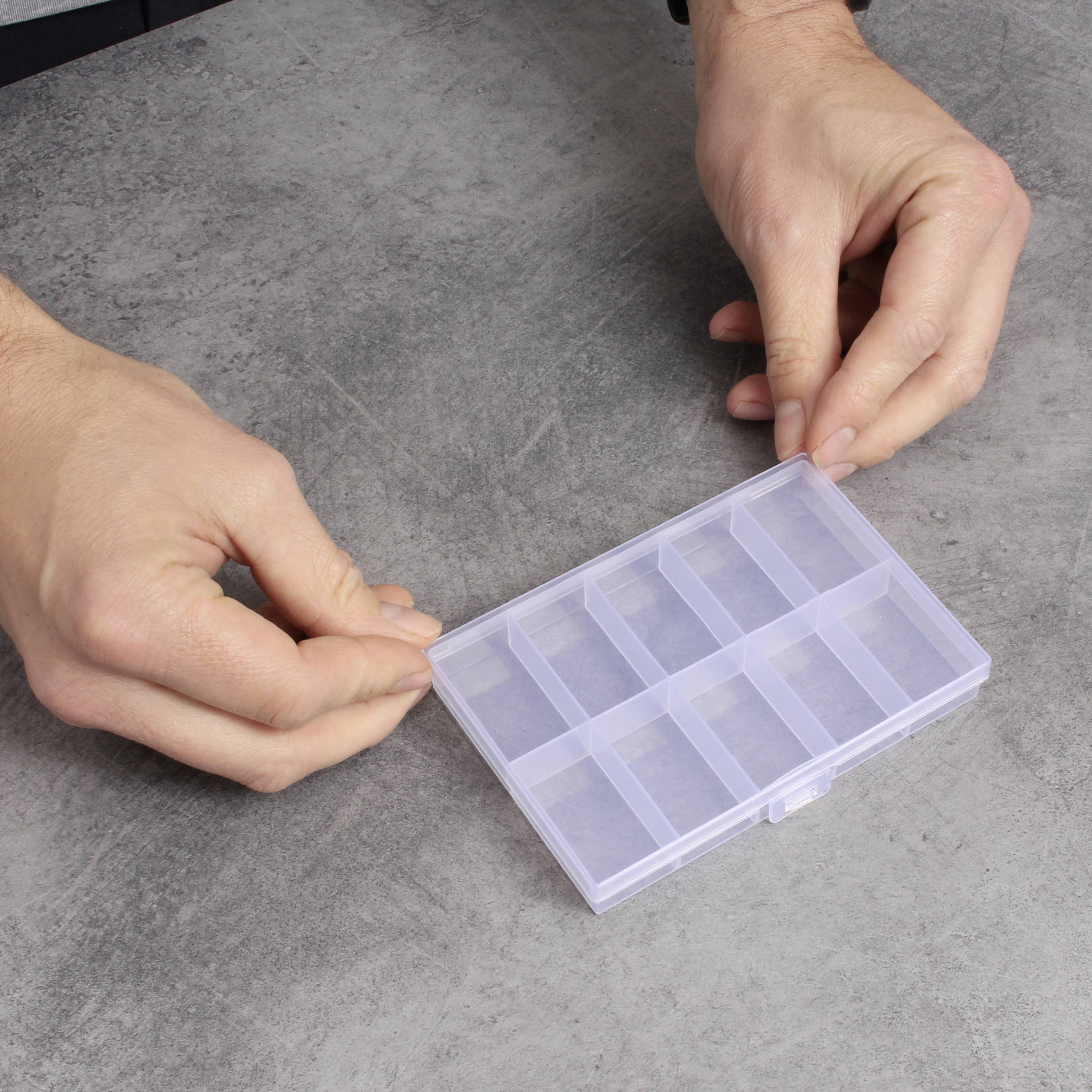 Osoba trzyma pustą, przezroczystą, prostokątną skrzynkę do przechowywania z wieloma przegródkami na szarym stole.