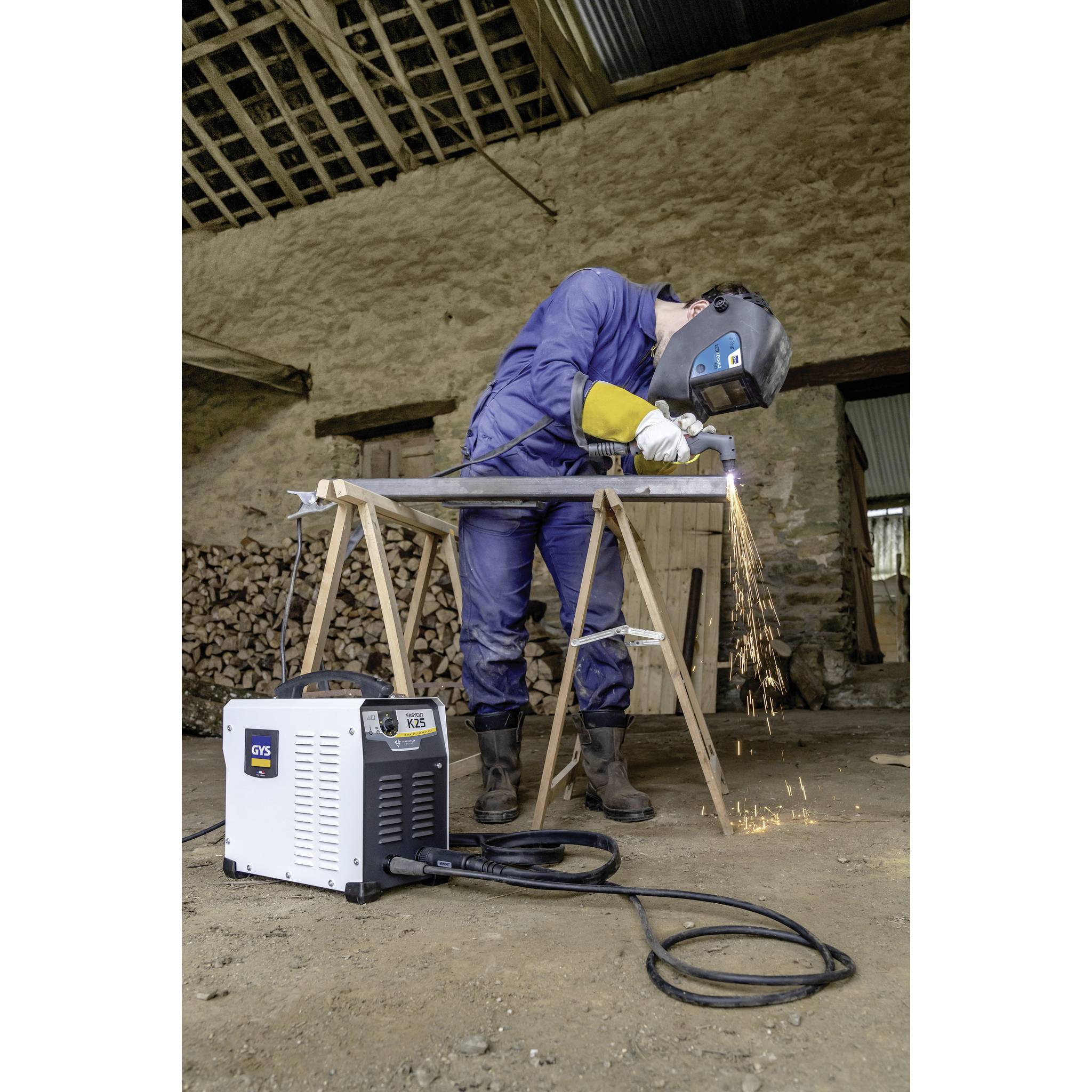 Osoba w odzieży ochronnej wykonuje prace spawalnicze na kawałku metalu na wolnym powietrzu. Lecą iskry. Sprzęt spawalniczy widoczny na pierwszym planie.