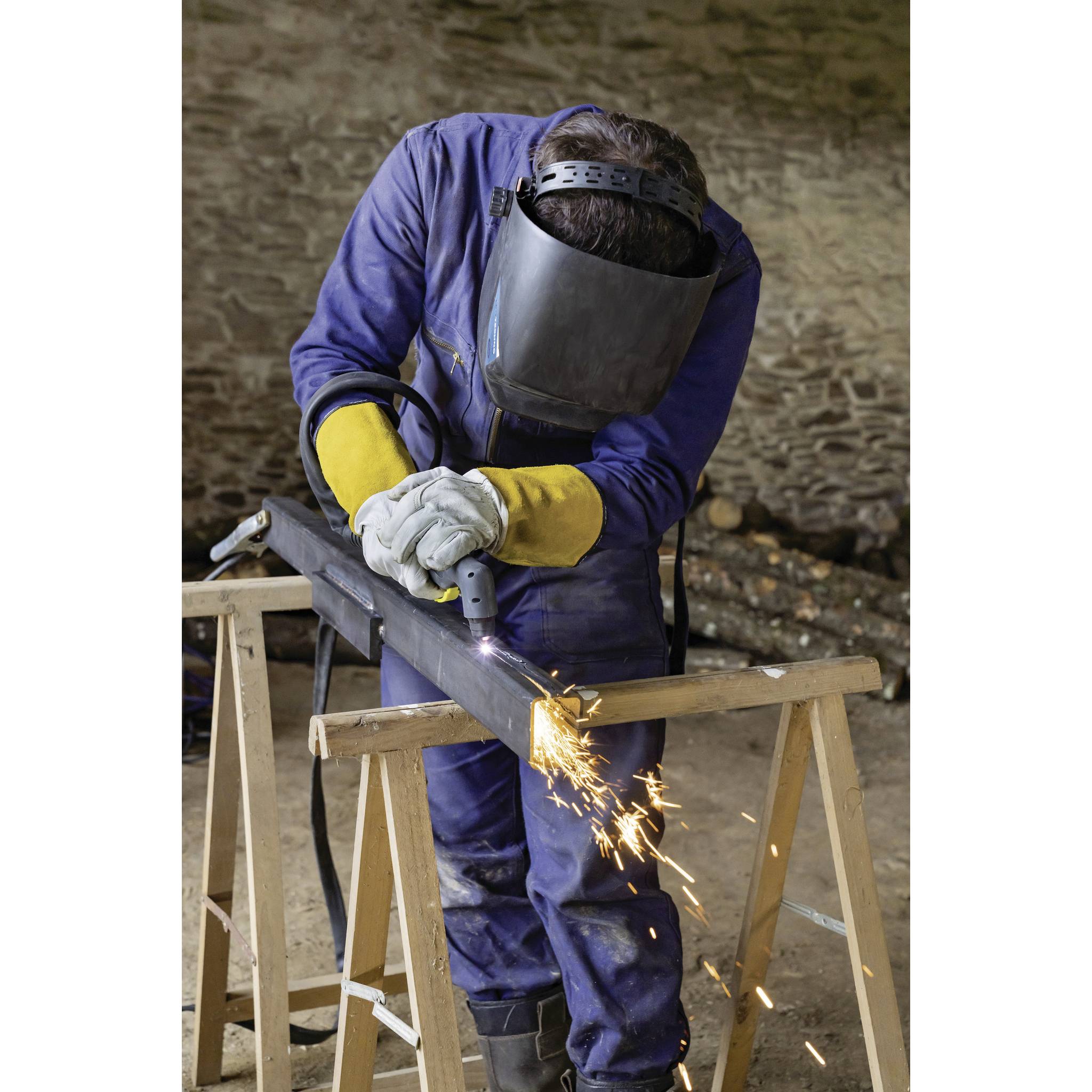 Osoba w masce ochronnej i rękawicach spawająca metal w warsztacie. Od spoiny odlatują iskry.