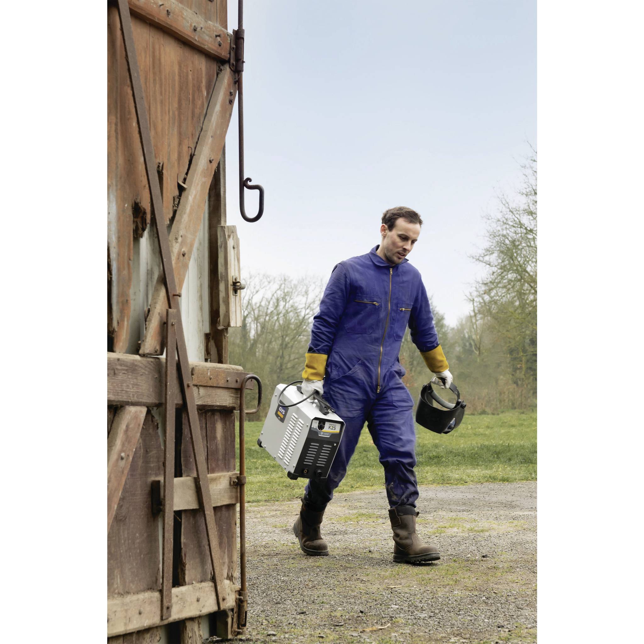 Mężczyzna nosi kask spawalniczy i trzyma w ręku sprzęt spawalniczy, stojąc na żwirowej drodze obok drewnianej bramy stodoły.