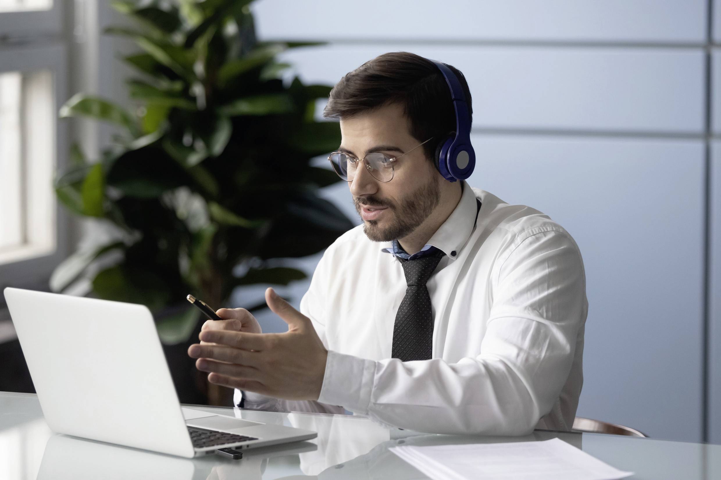 Mężczyzna w formalnej koszuli i krawacie siedzi przy biurku, ma słuchawki na uszach i mówi do mikrofonu. Przed nim leży laptop.