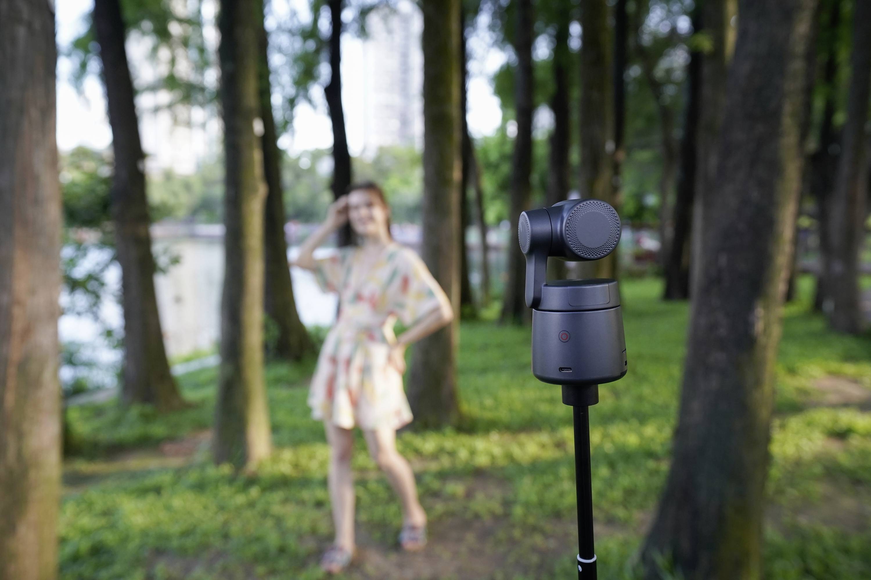 Kobieta pozuje w parku nad rzeką. Na pierwszym planie widać aparat na statywie skierowany w jej stronę.