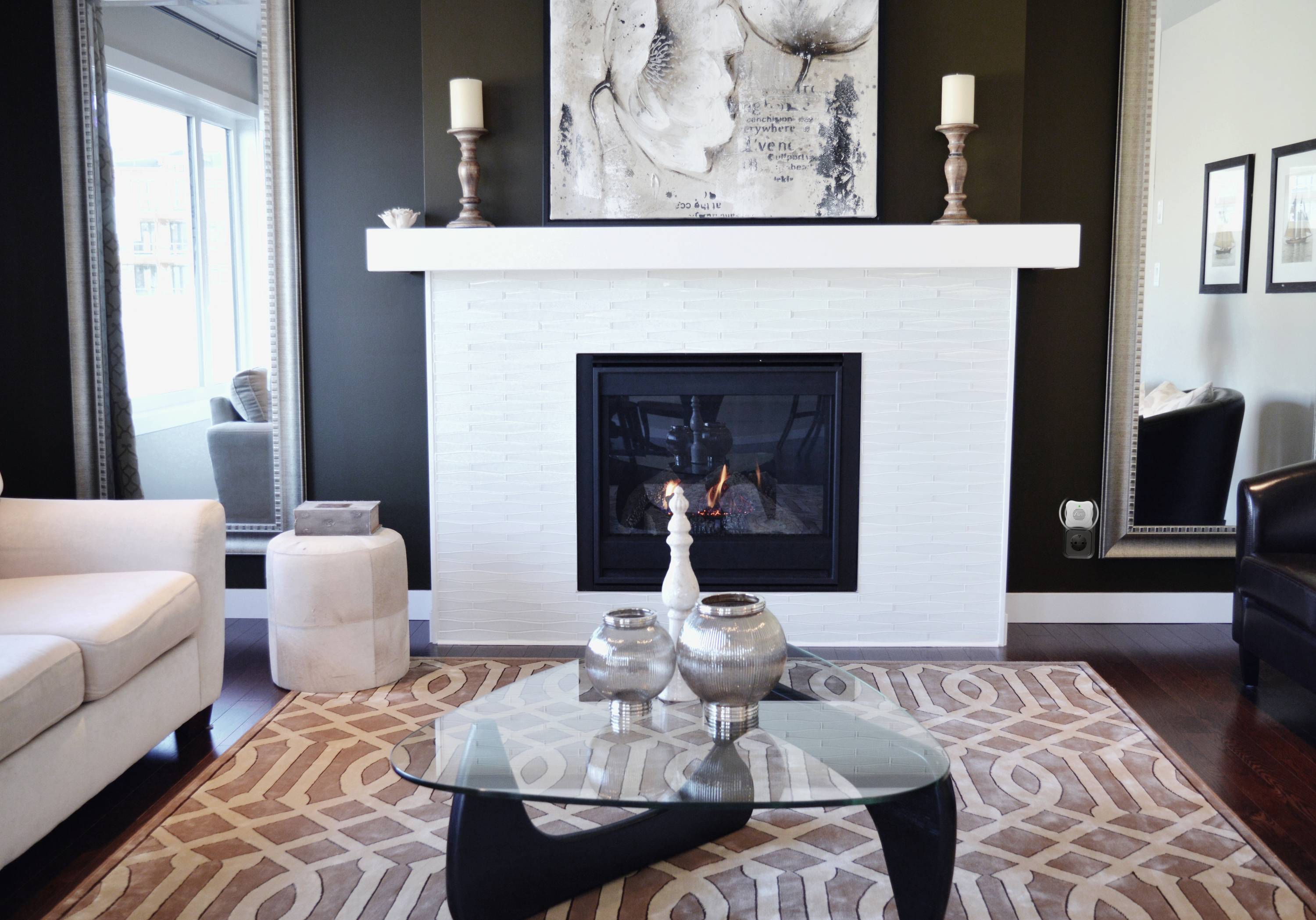 Salon z nowoczesnym kominkiem. Biała ceglana obudowa, powyżej dzieło sztuki. Szklany stolik kawowy z dekoracjami przed nim.